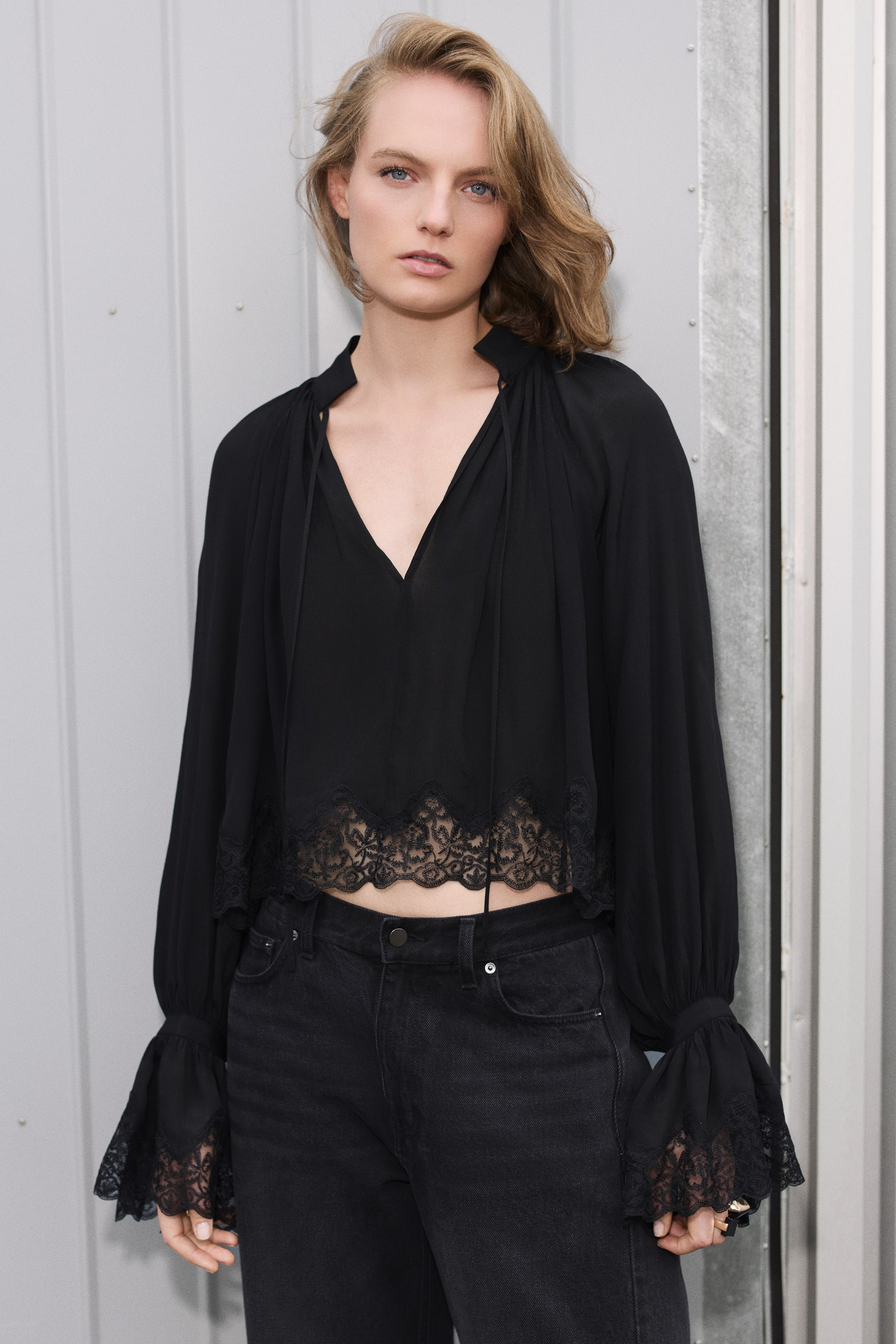 Ver imagen más grande: La modelo lleva una blusa negra de corte holgado y largo cropped, combinada con unos vaqueros negros. La blusa presenta cuello mao con amplio escote en V y largos lazos, mangas abullonadas con puños acampanados y ribete de encaje con acabado de ondas en los puños y el bajo.