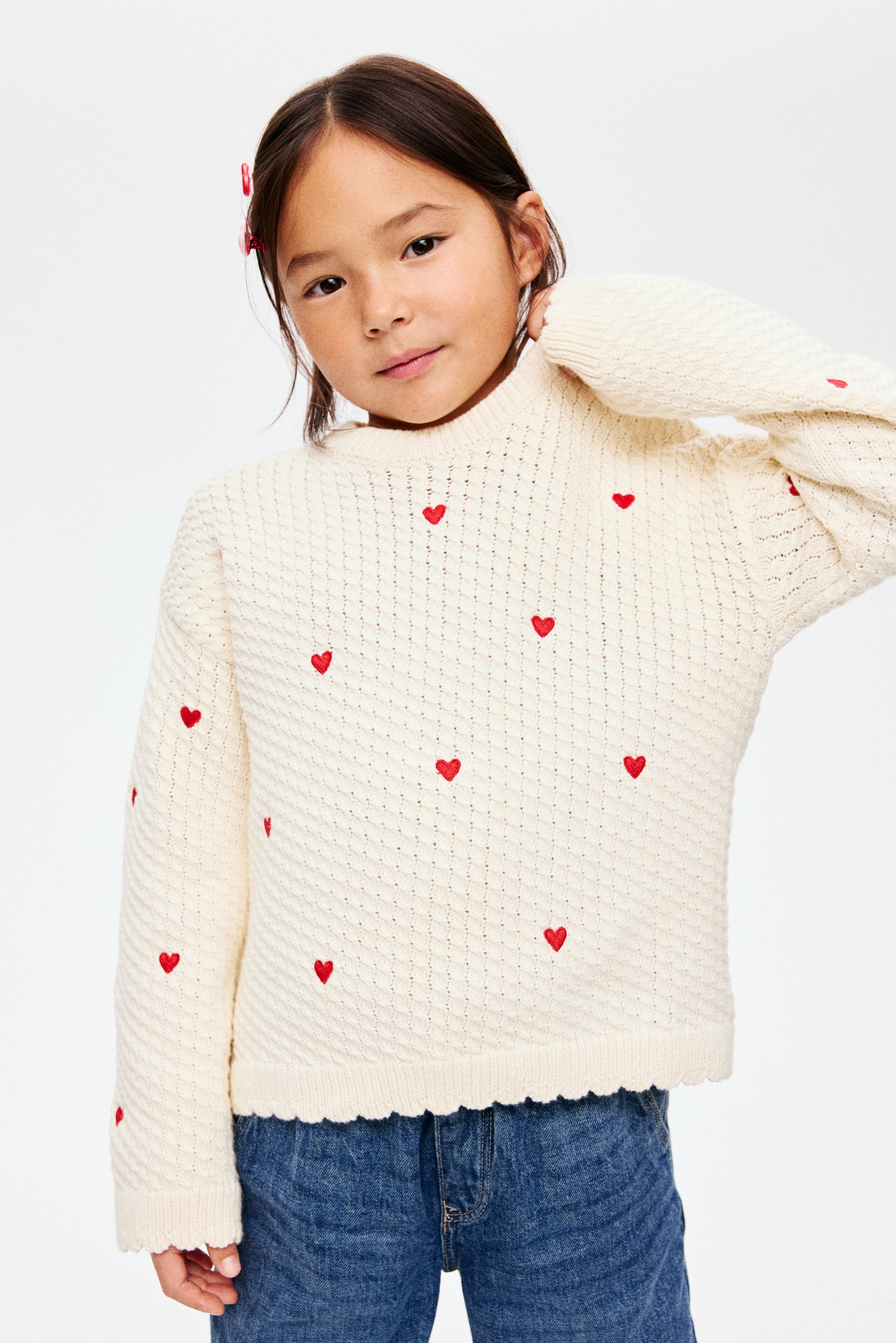 View larger image: A cream knit sweater is featuring a textured stitch pattern across its FRONT. It is displaying small red embroidered hearts scattered throughout, a mock neck, long sleeves, and scalloped hem and cuffs.