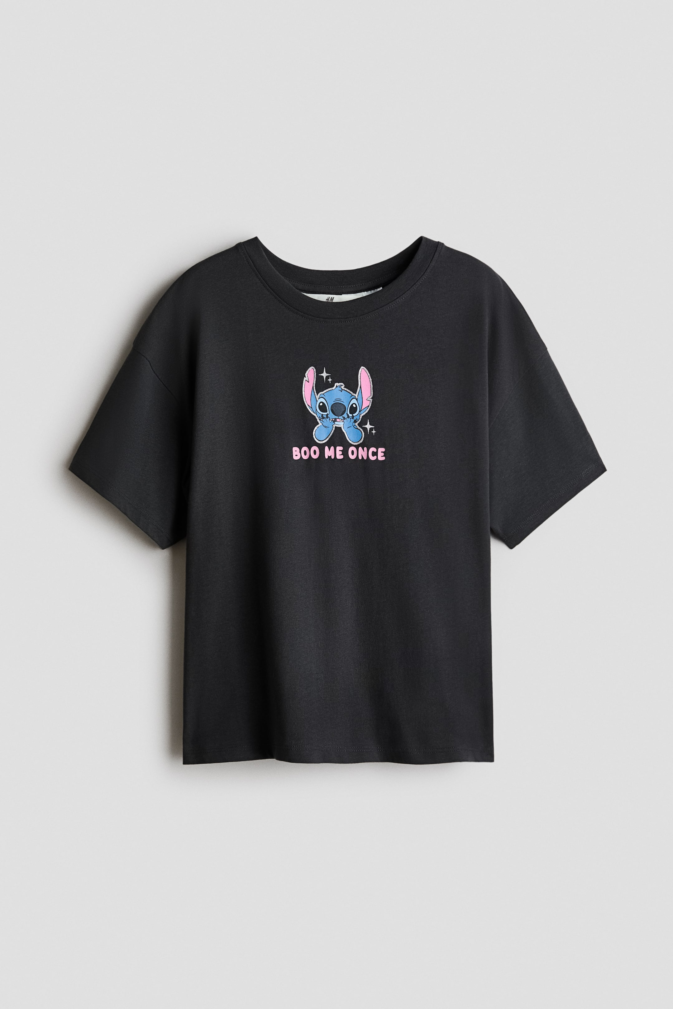 View larger image: A black short-sleeved crew neck T-shirt is laid flat, facing forward. It features a blue cartoon character with pink ears and small white star details on the front, and the text 'BOO ME ONCE' is printed in pink below the character.