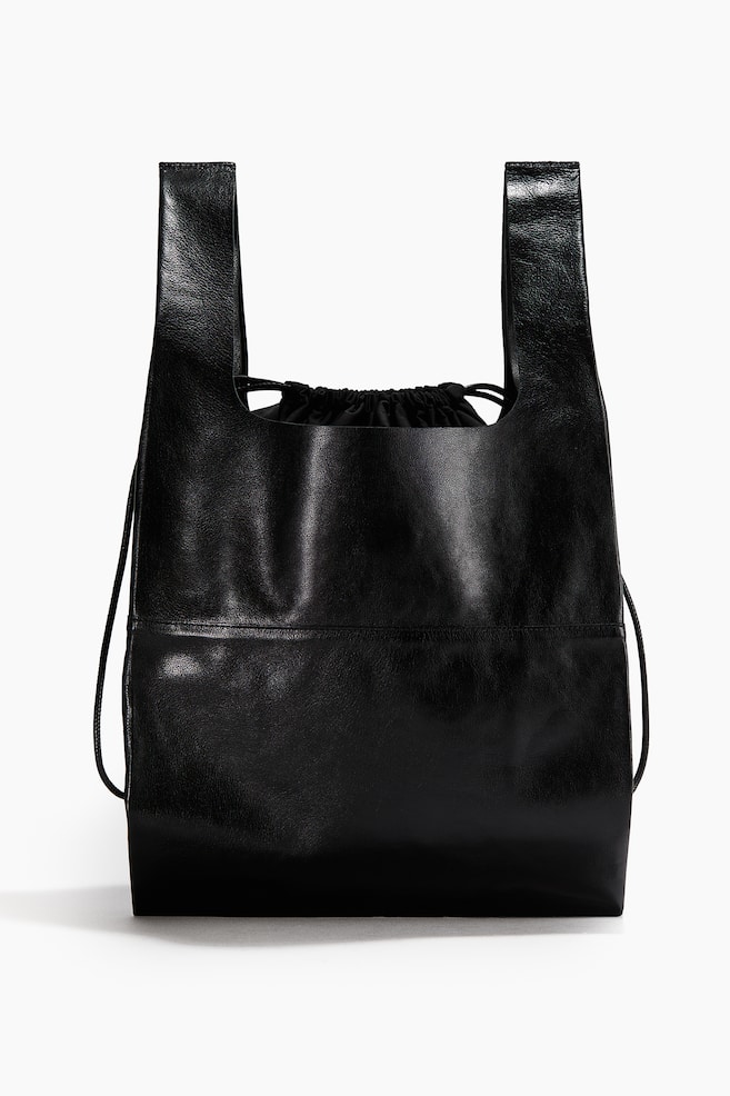 A black tote bag crafted from soft leather is standing front-facing, featuring two top handles with raw edges. A horizontal seam is visible across the front, and an inner black twill pouch with a drawstring closure is peeking from the top opening. - 1