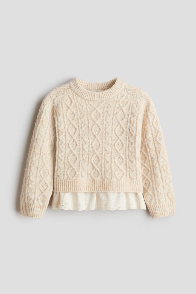 A cream-coloured cable-knit sweater is facing front, showcasing a ribbed crewneck, ribbed cuffs, and a ribbed hem. A white ruffled eyelet-lace underlayer is peeking out from beneath the bottom hem, creating a layered appearance. - 1