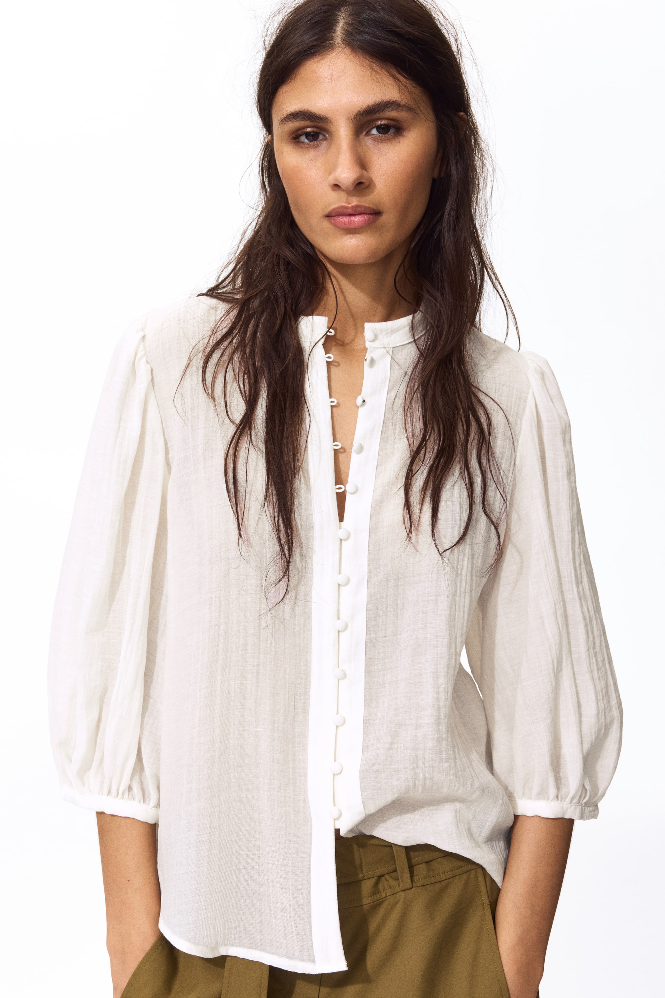 View larger image: A white textured blouse features a mandarin collar and a central button-up placket with small white buttons, some of which are undone to create a V-neck opening. It is designed with puffed three-quarter sleeves ending in gathered cuffs, and its curved hem rests untucked over olive green trousers.