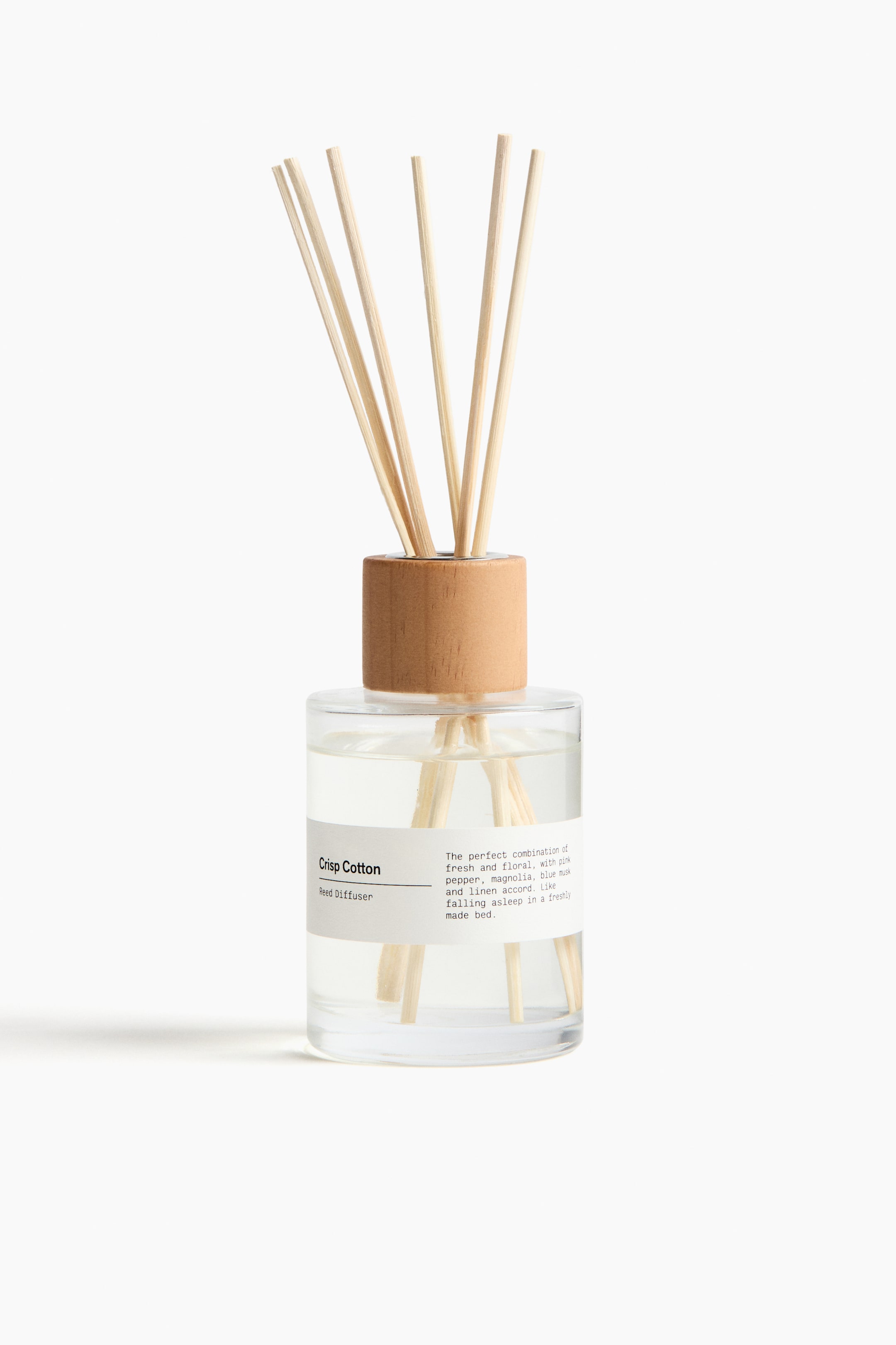View larger image: A reed diffuser is presenting front, showing a clear glass bottle holding a liquid and several light-coloured wooden diffuser sticks standing upright inside. A pinewood ring is encircling the bottle's opening, and a white label reading "Crisp Cotton Reed Diffuser" is adhering to the front.