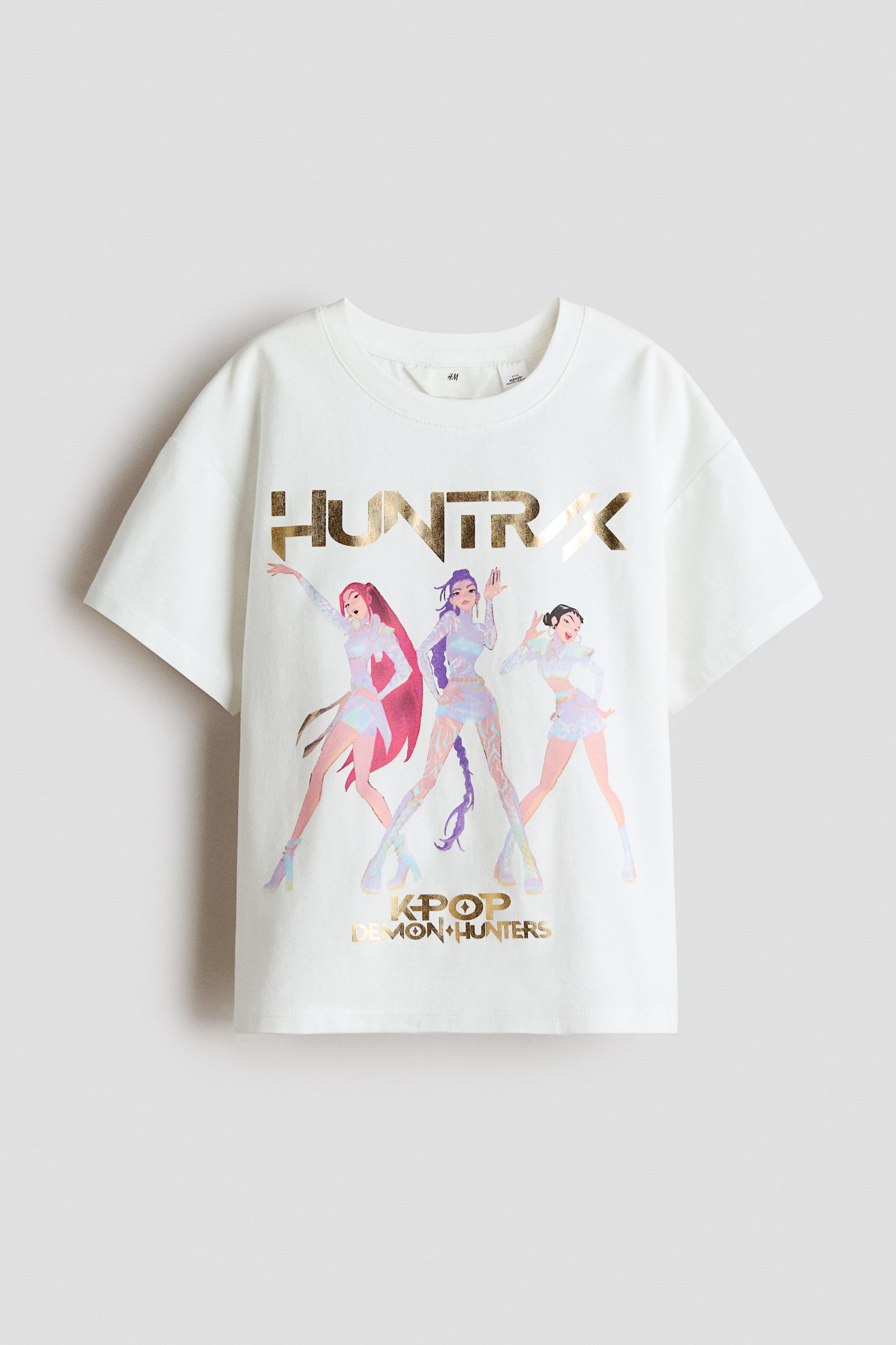 Ingrandisci l'immagine: Una t-shirt oversize è mostrata di fronte, caratterizzata da uno scollo rotondo con bordo a coste e spalle cadenti. La t-shirt color crema presenta una grande stampa grafica con la scritta 'HUNTRX' in lettere dorate metallizzate sopra tre figure femminili stilizzate in colori iridescenti, e la frase 'KPOP DEMON HUNTERS' in lettere dorate metallizzate abbinate sotto.