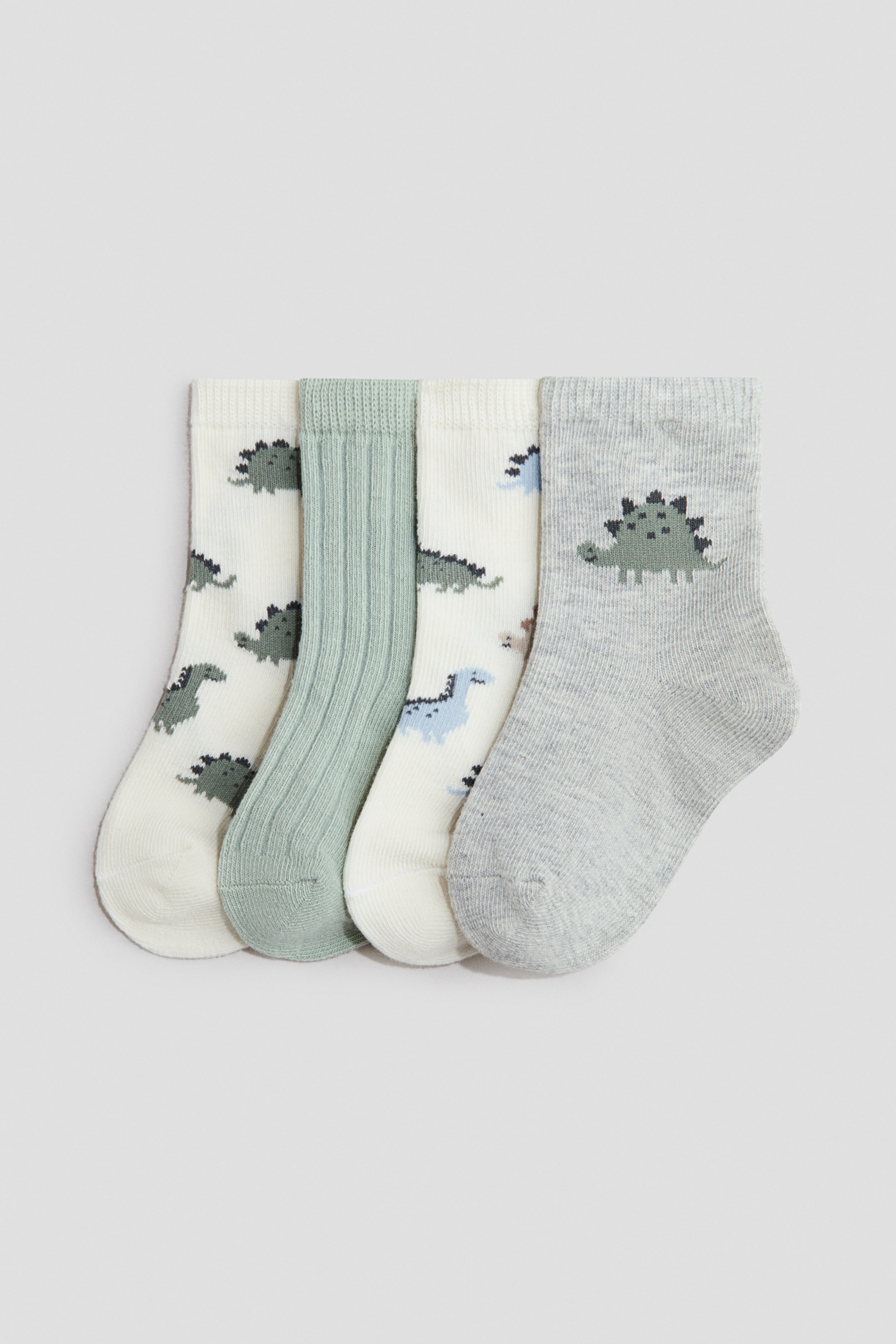 View larger image: A set of four socks is showcasing their front sides, arranged side-by-side. Two cream socks feature pixelated dinosaur patterns, one with dark green dinosaurs and another with light blue and brown dinosaurs. A light green sock displays a ribbed texture, and a light grey sock shows a large dark green pixelated dinosaur on its front.