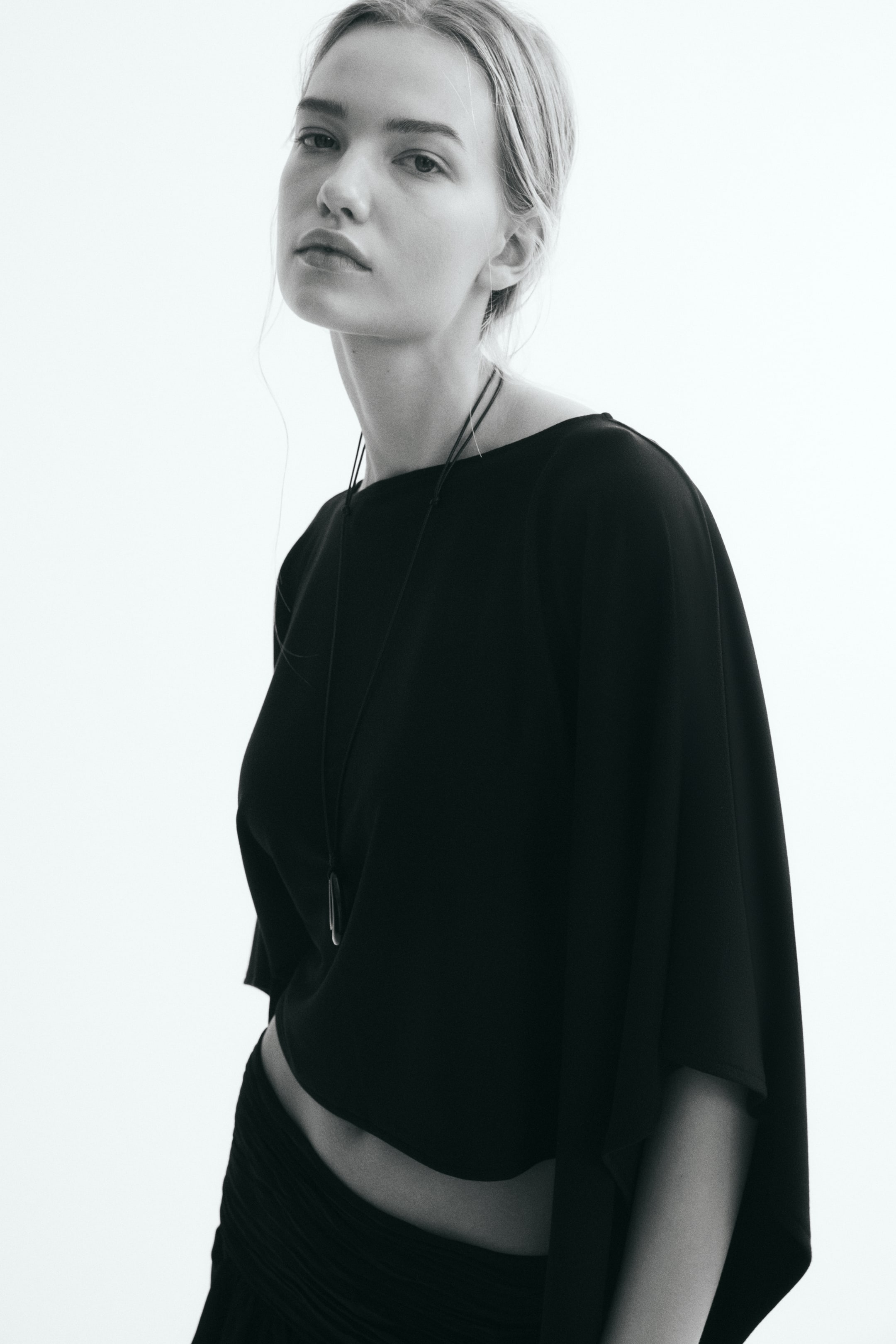 View larger image: A short, loose-fit black cape featuring a boat neckline and a draped, asymmetrical handkerchief hem is worn by the model. A thin cord necklace with a dark pendant is layered over the front of the cape.