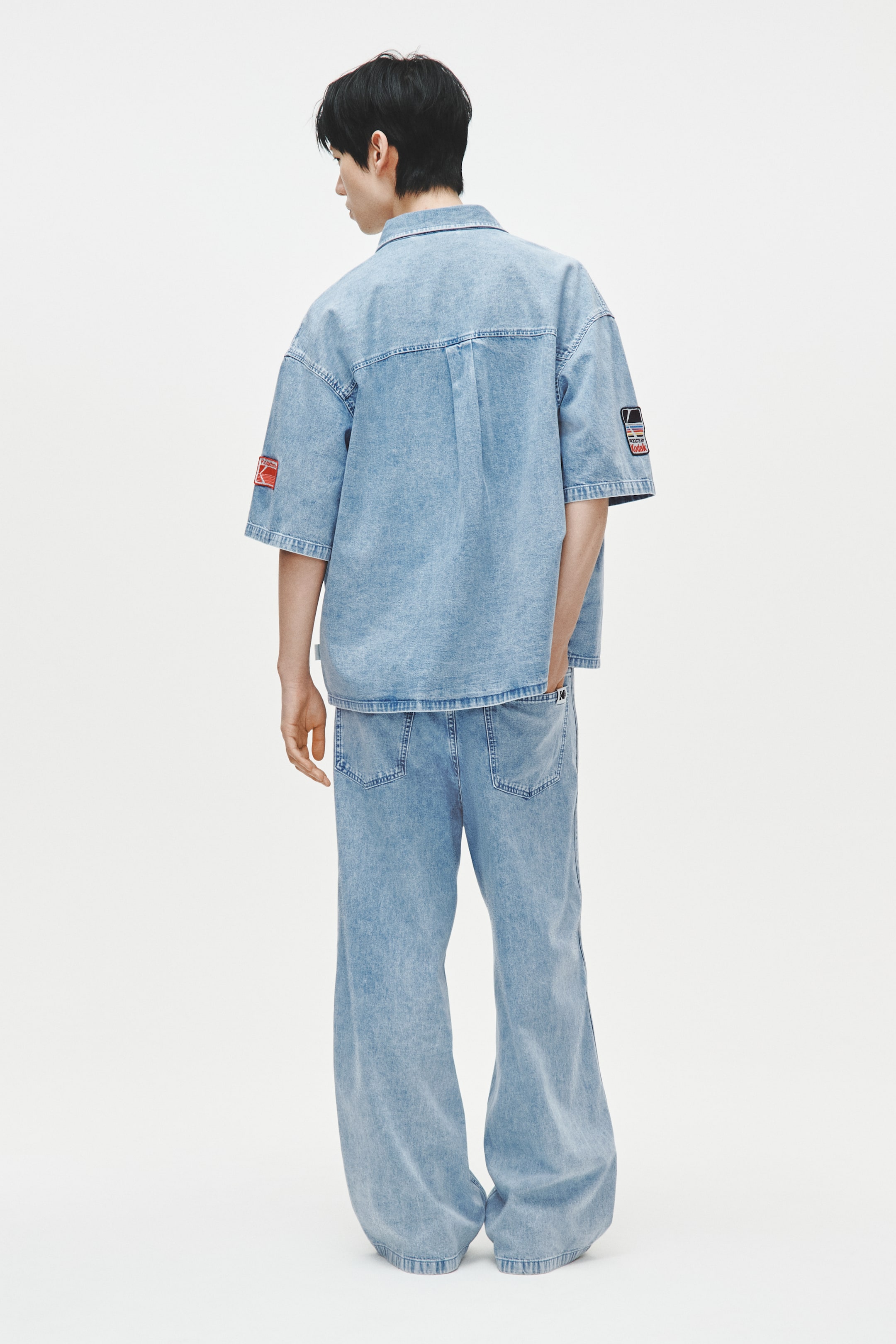 View larger image: "Isang light blue na denim short-sleeved shirt at katugmang wide-leg denim pants ang suot, tinitingnan mula sa likod. Ang oversized na shirt ay may back yoke na may pleats at isang pulang rectangular patch sa kaliwang manggas at isang multi-colored graphic patch sa kanang manggas, habang ang pantalon ay nagtitipon sa mga bukung-bukong."