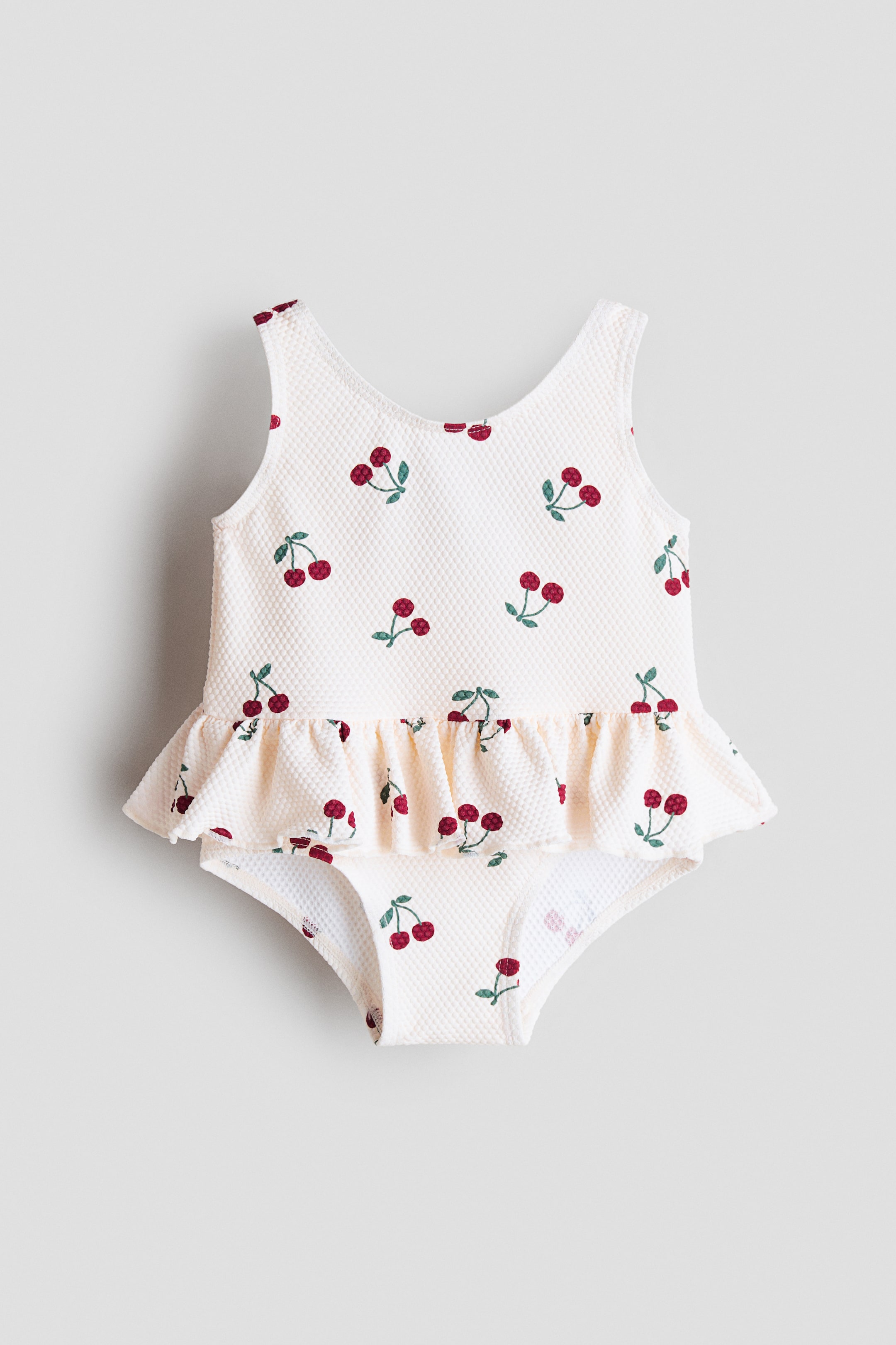 View larger image: A textured swimsuit, front-facing, features a round neckline, a decorative flounce at the waist and a lined gusset. It has an all-over red cherry pattern on a white background.