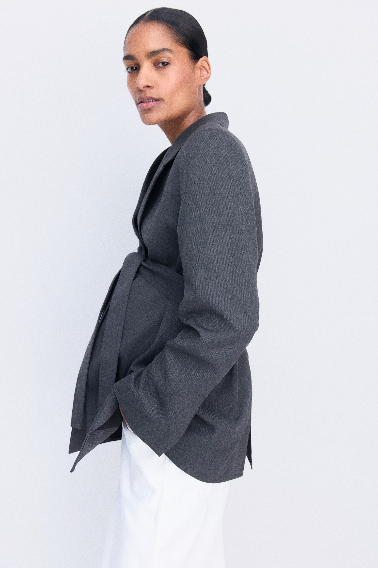 A dark grey woven blazer featuring notch lapels and a matching self-tie belt at the waist is being worn by the model. The blazer is styled with white trousers, and the model is posing in a side profile view. - 1