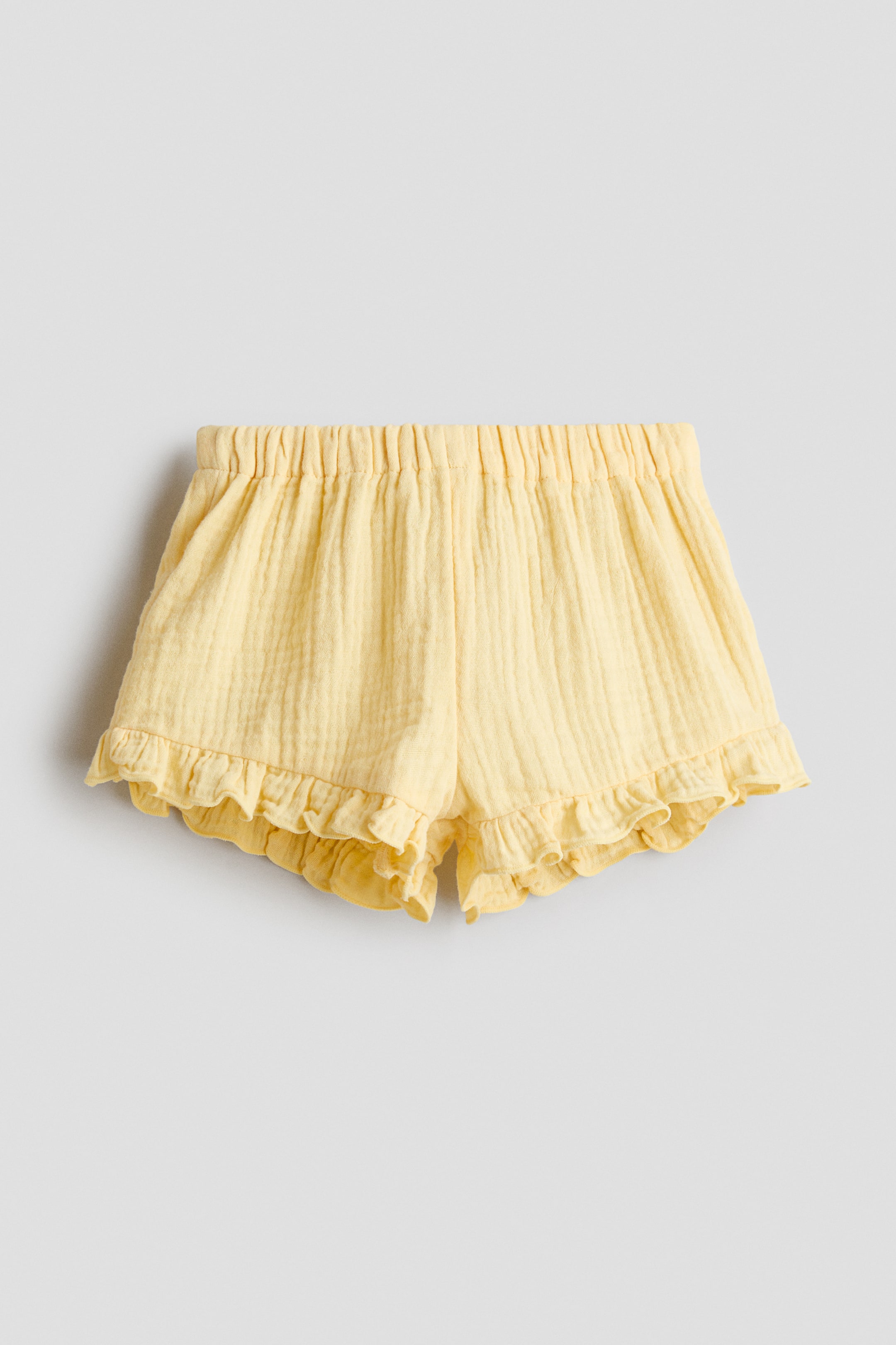 View larger image: Yellow shorts feature a crinkled texture, an elasticated and gathered waistband, and ruffled hems. They display a loose, short fit from the front.