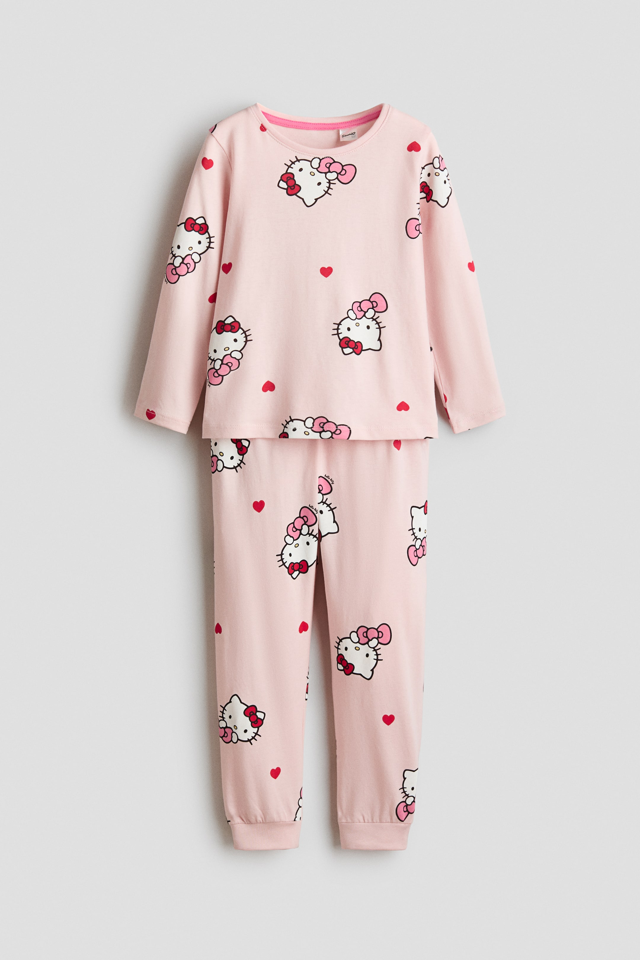 Näytä suurempi kuva: Vaaleanpunainen kaksiosainen pyjamaesittely on kuvattu tasokuvaan aseteltuna, ja siinä on toistuva Hello Kitty -kasvojen kuvio vaaleanpunaisilla ruseteilla ja pienillä punaisilla sydämillä. Pitkähihainen yläosa on pyöreäaukkoista mallia, ja pitkät housut päättyvät joustaviin nilkkalahkeisiin.