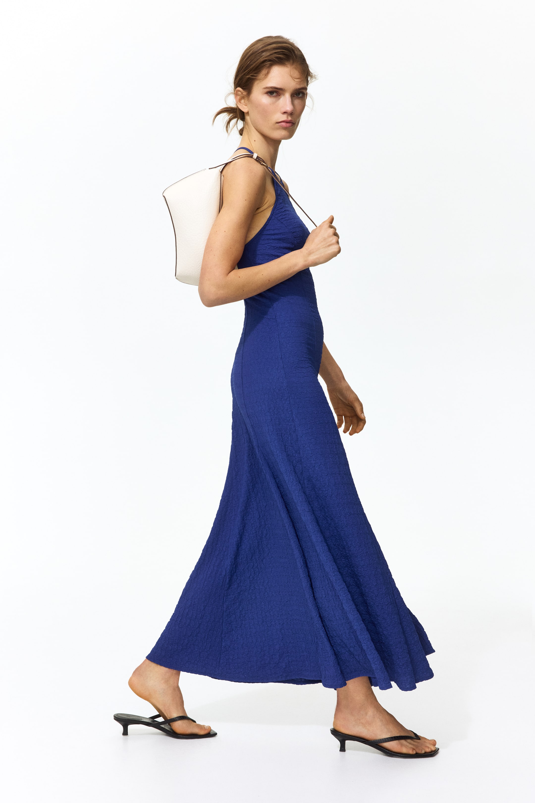 View larger image: A royal blue textured maxi dress with thin shoulder straps and a fitted bodice flows into a flared skirt, shown from a side angle. It is styled with a white shoulder bag and black low-heeled thong sandals.