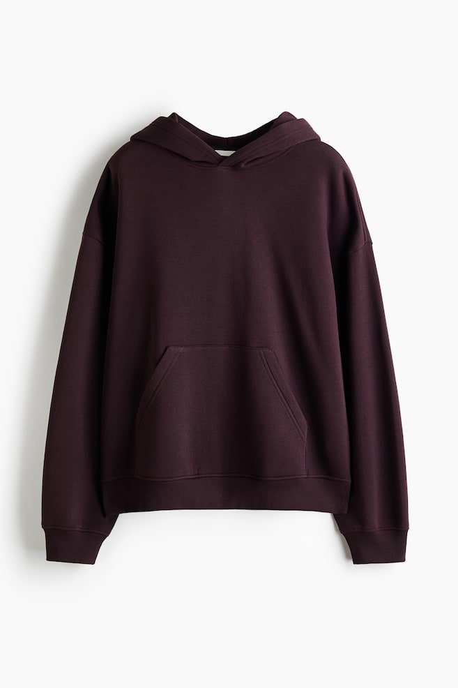 En mørk burgunder hoodie hænger med forsiden vendt fremad og fremhæver dens afslappede pasform med lave, sænkede skuldre og en dobbeltlaget hætte. Designet har en kængurulomme foran og er afsluttet med ribkanter ved ærmerne og en ribkant forneden. - 1