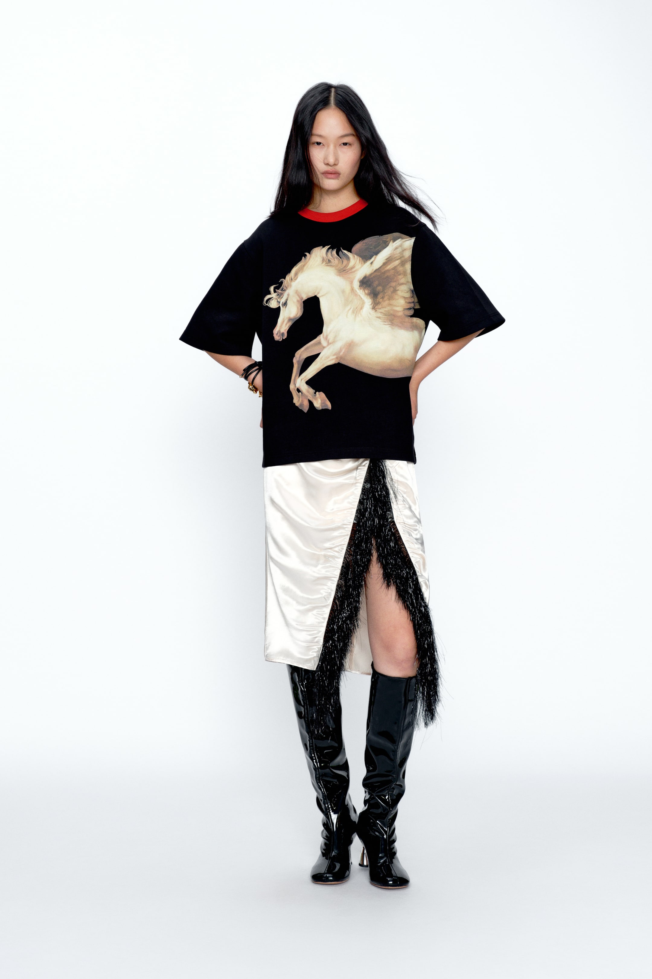 View larger image: A black oversized T-shirt featuring a large, light-toned pegasus graphic rearing on the front and a red crew neckline. It is styled over a cream satin skirt with a front slit trimmed in long black fringe, paired with black patent knee-high boots.