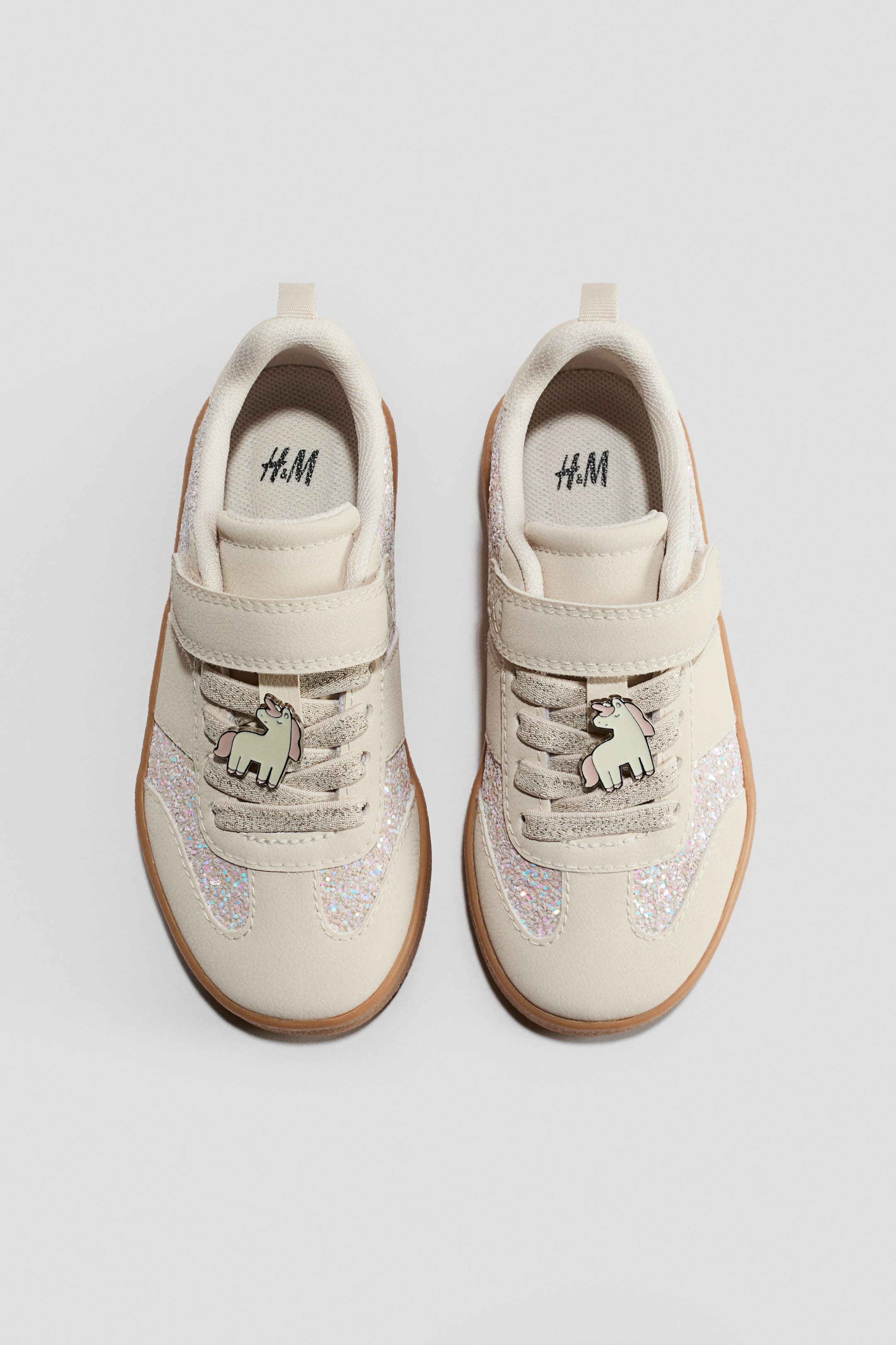 Grotere afbeelding bekijken: Trainers. Het FRONT-view toont een paar crèmekleurige lage trainers met glitterpanelen aan de zijkanten en op de neus. Elke trainer wordt vastgemaakt met één klittenbandriem over glinsterende veters, heeft een eenhoornbedel aan de veters en rust op een bruine rubberen zool.