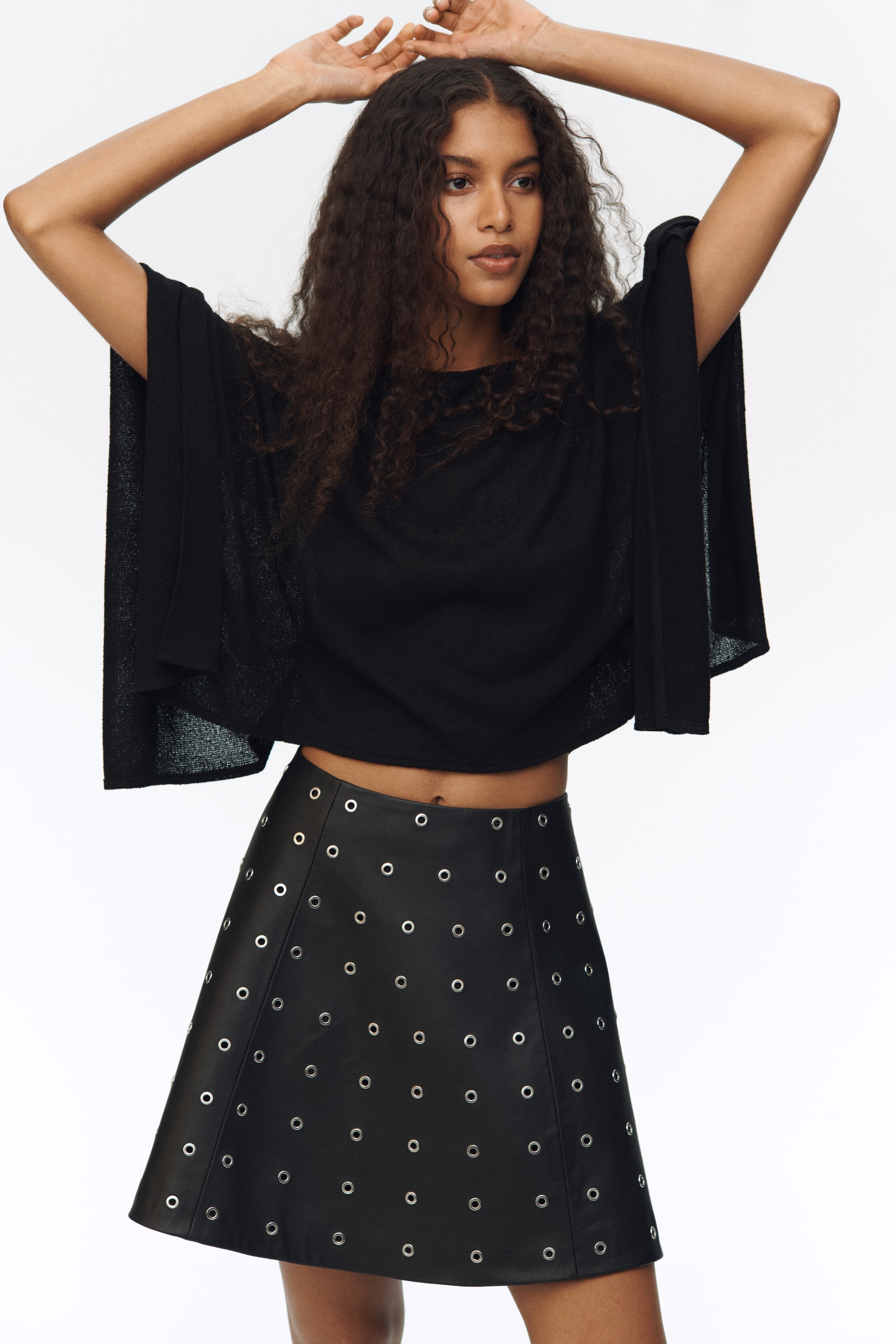 הצגת תמונה גדולה יותר: חצאית מיני שחורה מעור בגזרת A מוצגת מזווית קלה מלפנים-ימין, עם עיניות כסף רבות המסודרות בקווים אנכיים.