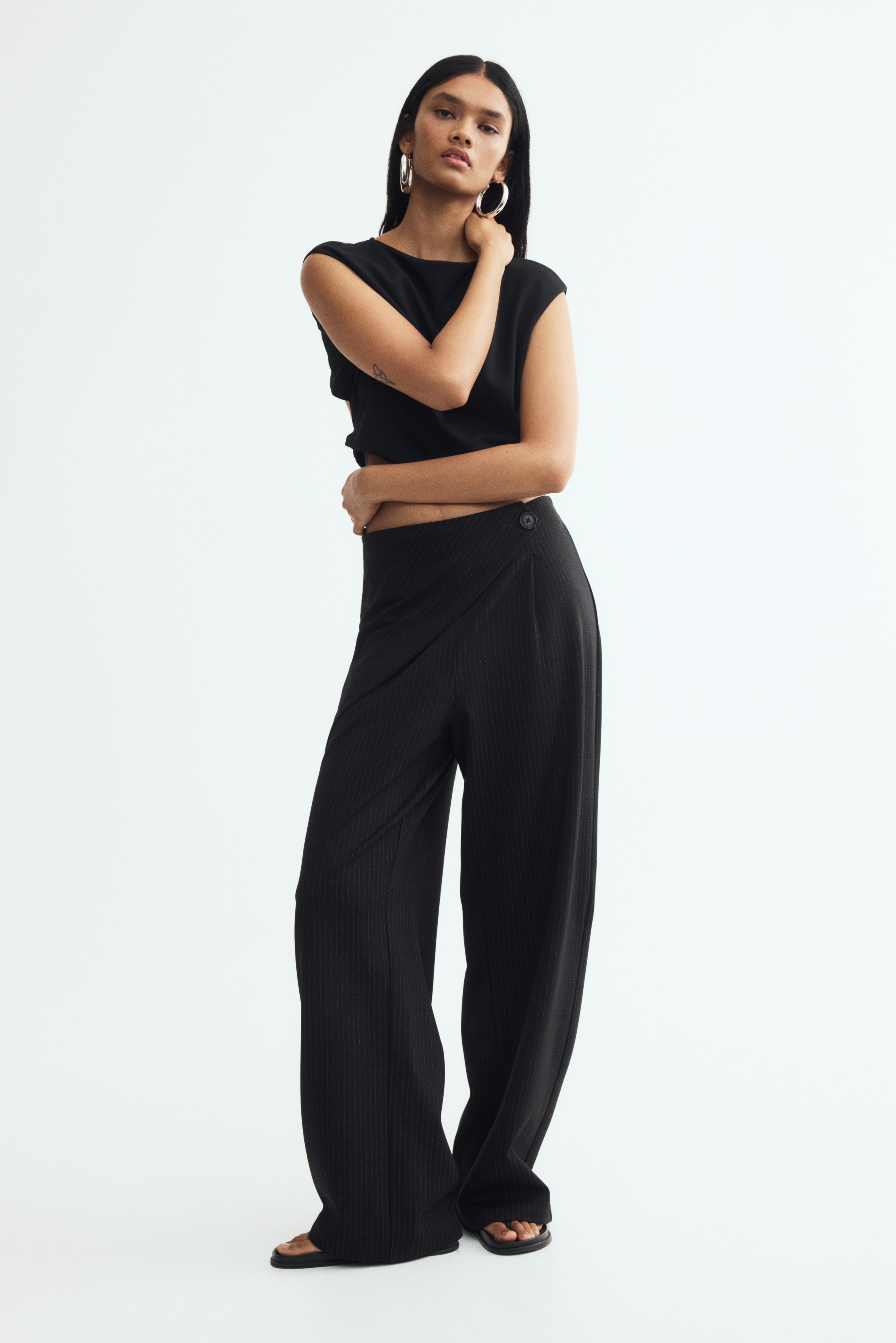 Zwarte broek met krijtstrepen met een voorzijde in wikkelmodel, waarbij diagonale krijtstrepen zichtbaar zijn op het overlappende pand. Een enkele zwarte knoop sluit de asymmetrische sluiting aan de rechterzijde, en de broek heeft een wijde pijp.