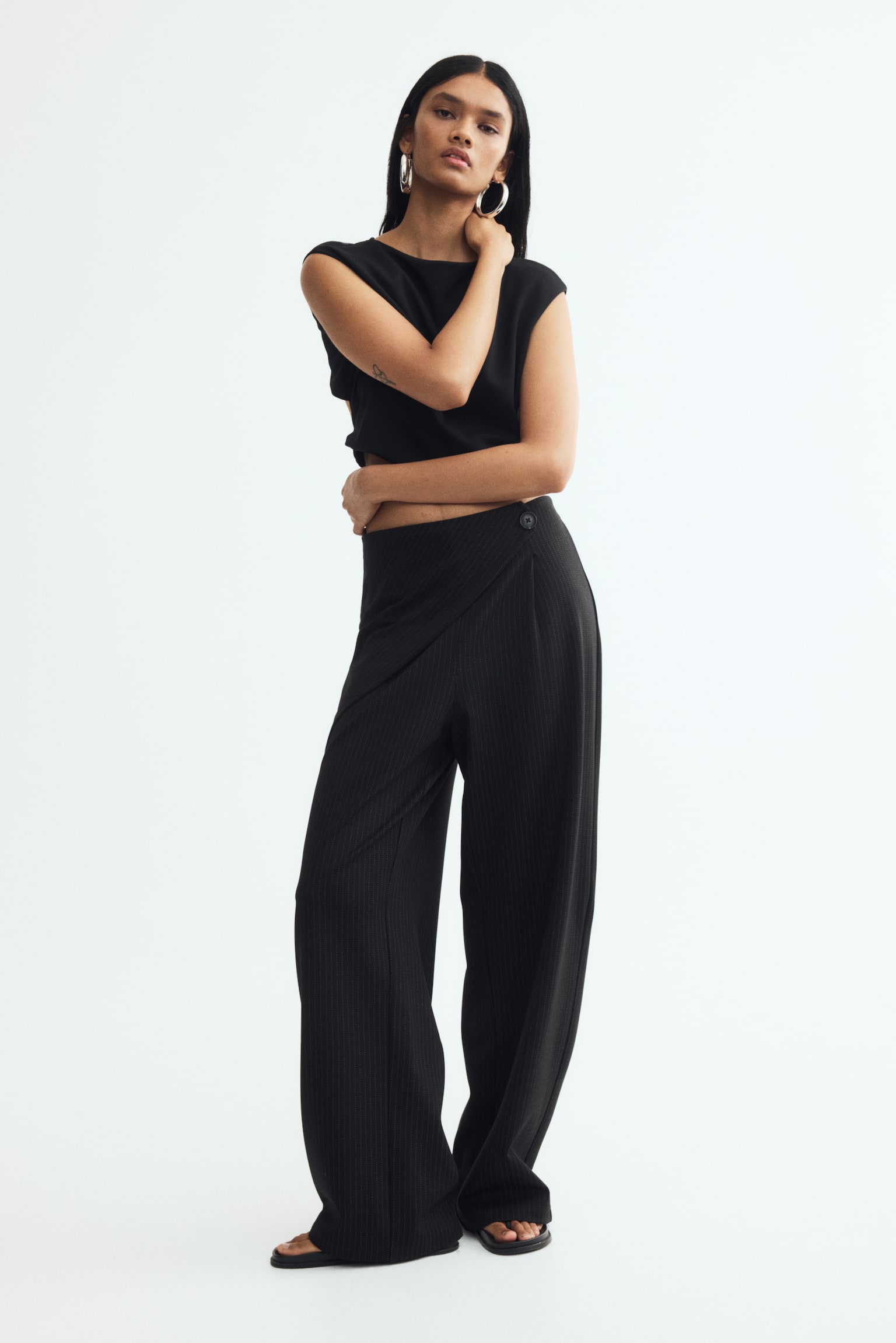 Black pinstriped trousers are displaying a front wrap-around design with diagonal pinstripes on the overlapping panel. A single black button is securing the asymmetrical closure on the right side, and the trousers are featuring a wide-leg cut. - 1