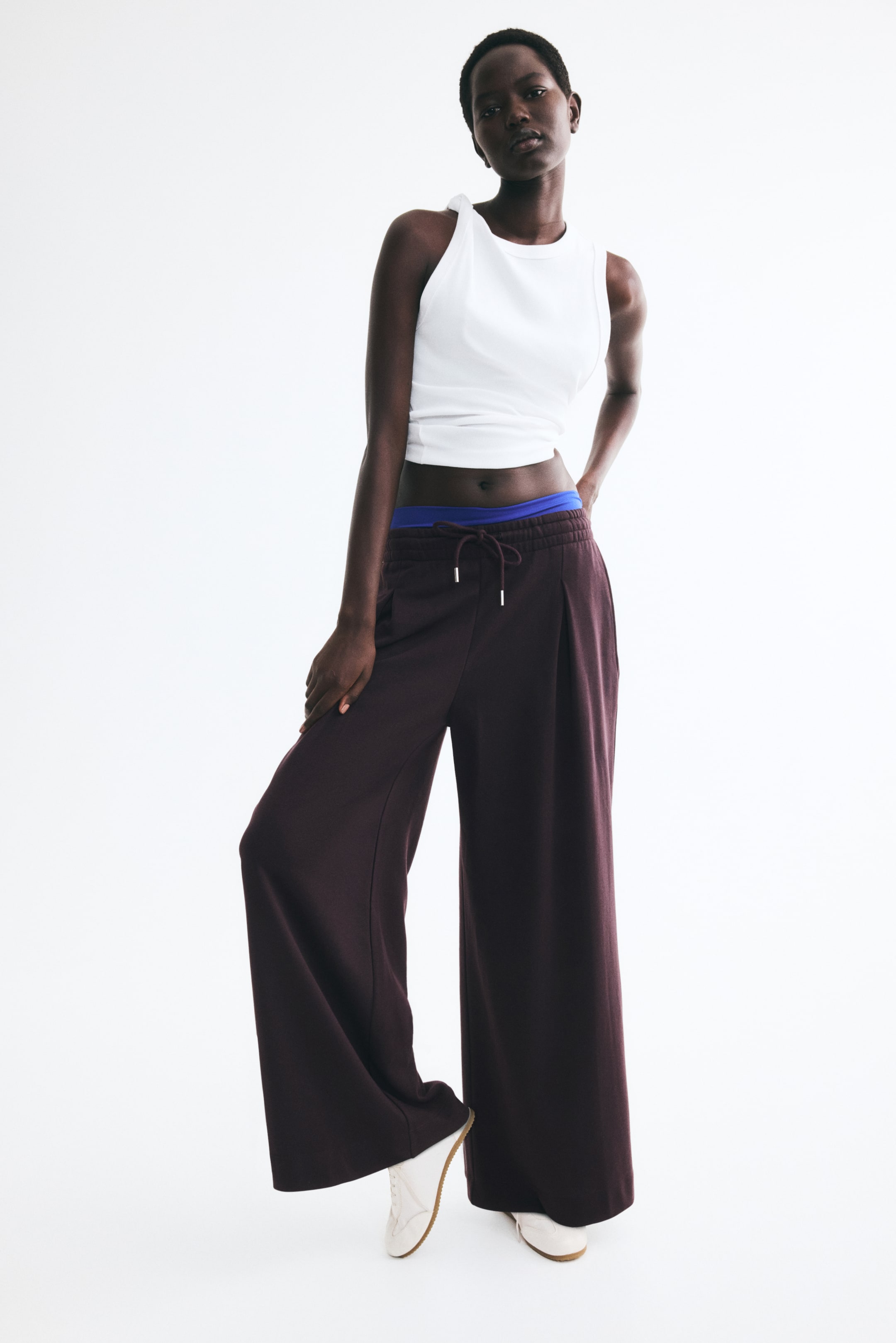 Visa större bild: Mörkt plommonfärgade sweatpants med lös passform, resårmidja med dragsko och vida, extra långa ben med inverterade boxveck bärs av modellen. Byxorna är stylade med ett vitt ärmlöst linne, vita sneakers och ett blått midjelager som syns ovanför midjan.