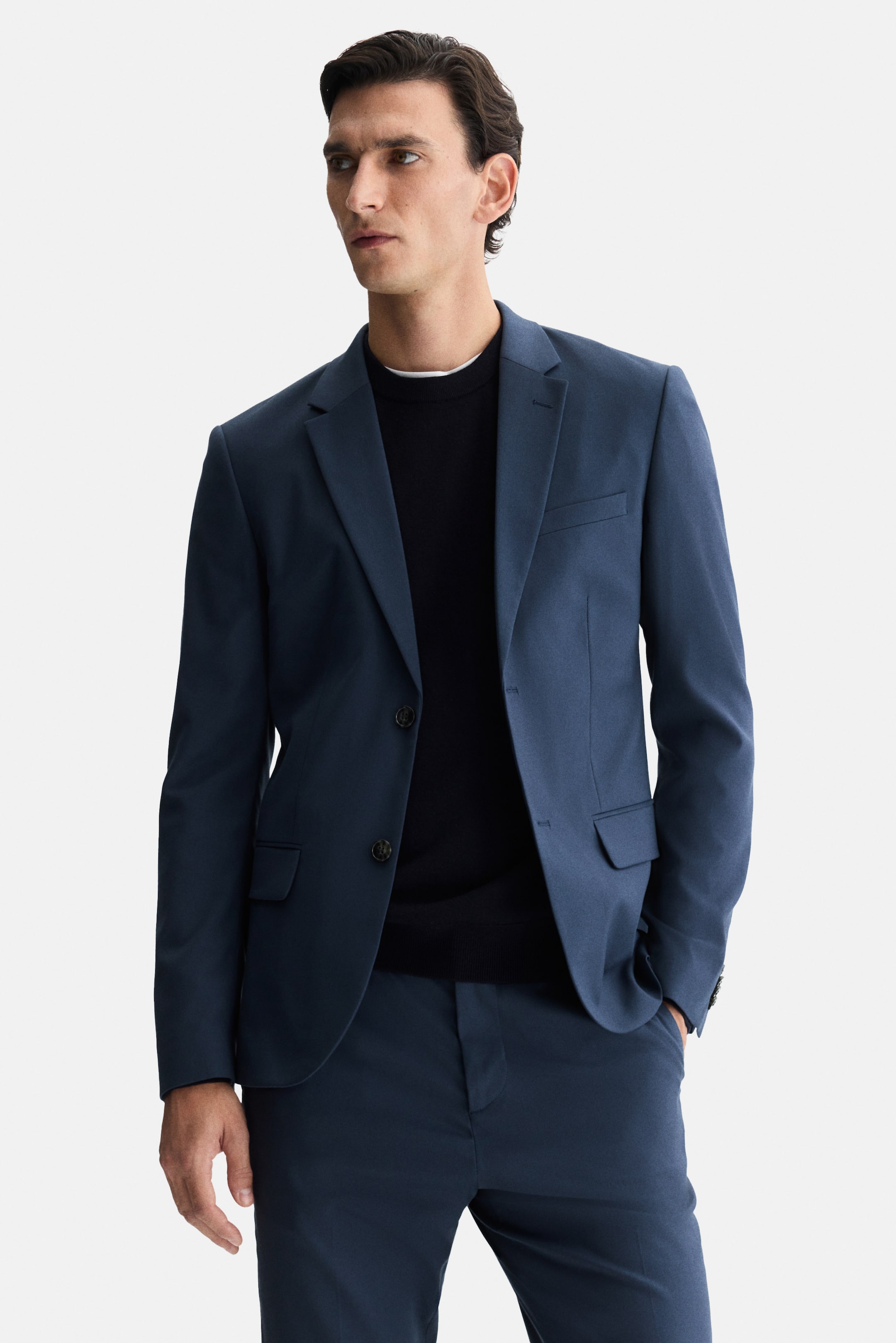 Ampliar la imagen: Un traje azul índigo, que consiste en un saco de un solo botón y pantalones, está siendo usado por el modelo. La chaqueta se lleva abierta sobre un suéter oscuro de cuello redondo, y una de las manos del modelo está siendo colocada en un bolsillo del pantalón.