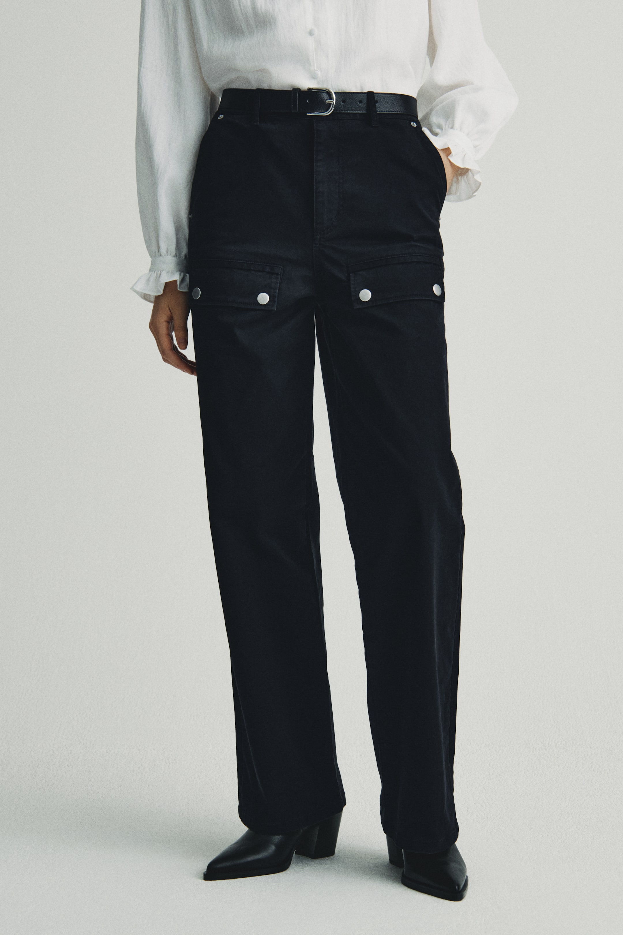 Xem ảnh lớn hơn: Quần ống rộng màu đen đang được trình bày từ phía trước, hơi nghiêng, cho thấy thiết kế cạp cao được thắt chặt bởi một chiếc thắt lưng đen. Hai túi nắp, mỗi túi được cố định bằng hai nút bấm màu bạc, có thể thấy ở phần đùi trên, và quần rơi thẳng, rộng, che phủ giày bốt mũi nhọn màu đen.