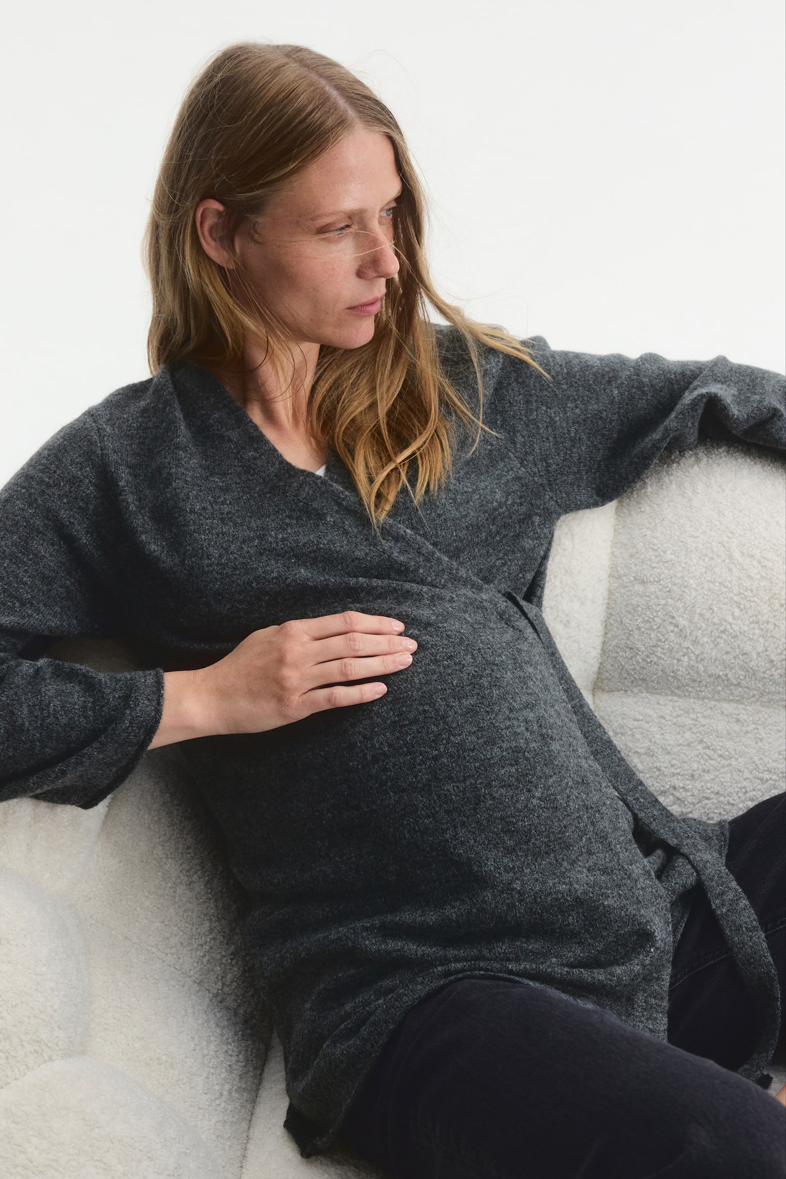 A dark grey, fine-knit wrapover cardigan is being worn by the model, who is resting one hand on their pregnant stomach. The cardigan features dropped shoulders, a V-neckline, and a soft texture, and is paired with dark trousers. - 1