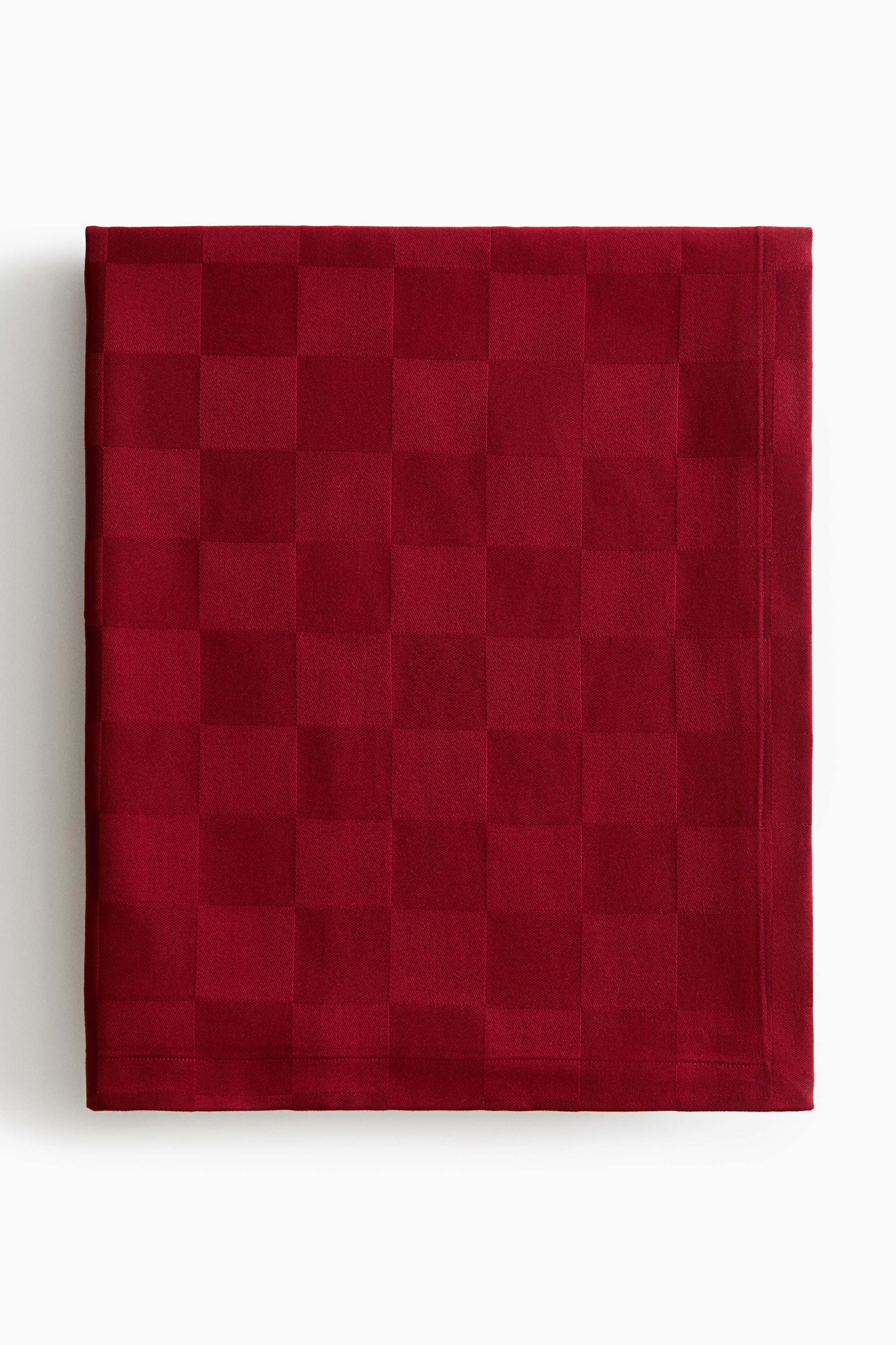 Ver imagen más grande: Se muestra de frente un mantel rojo oscuro plegado con un estampado de tablero de ajedrez en tono sobre tono, que presenta cuadrados alternos de texturas mate y lustrosas, rematado con bordes cuidadosamente dobladillados.