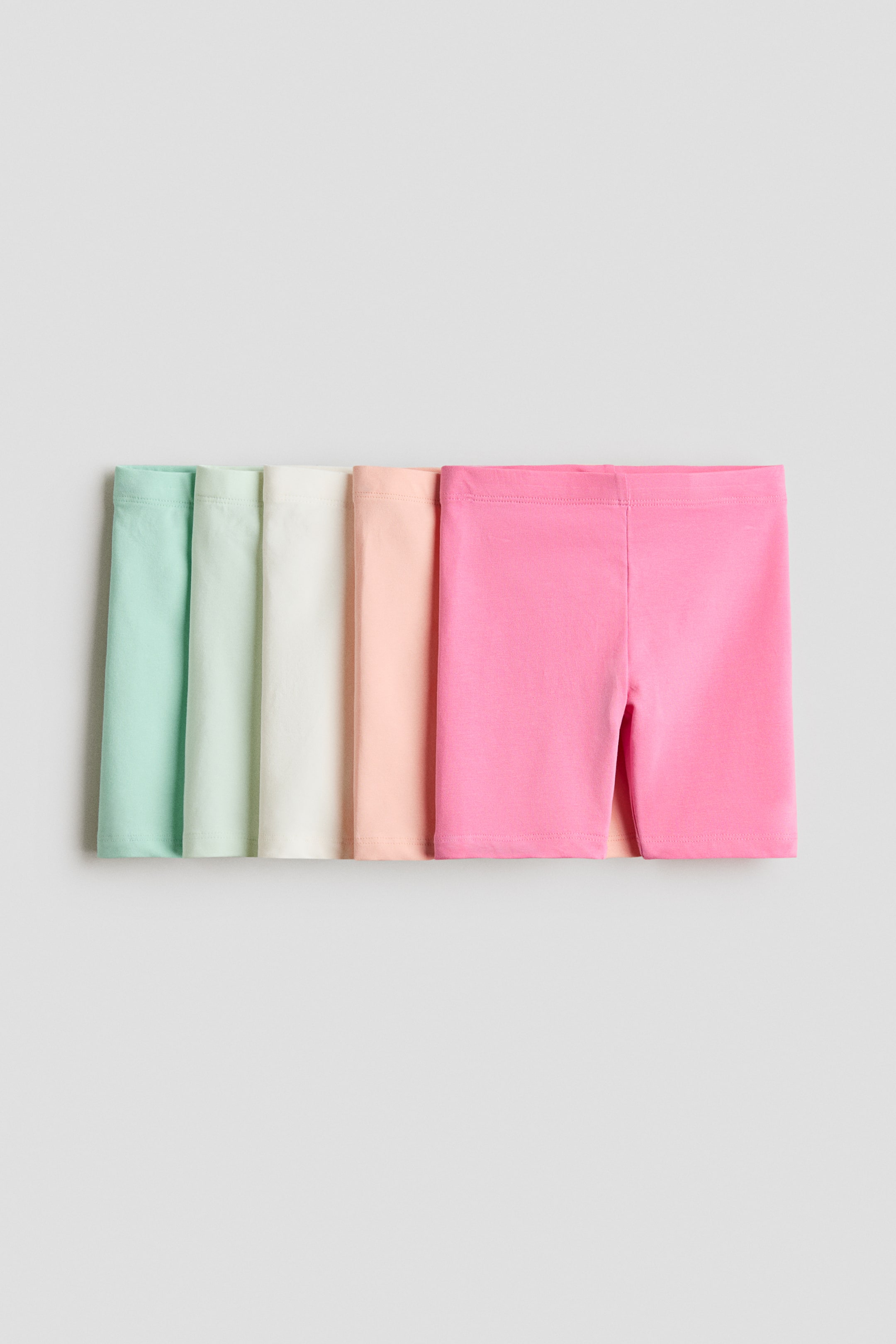 View larger image: A stack of cycling shorts in cotton jersey is showcased in five different colors. The front of the pink pair is fully visible, highlighting an elasticated waist, while mint green, light green, cream, and light peach shorts are partially visible behind it.
