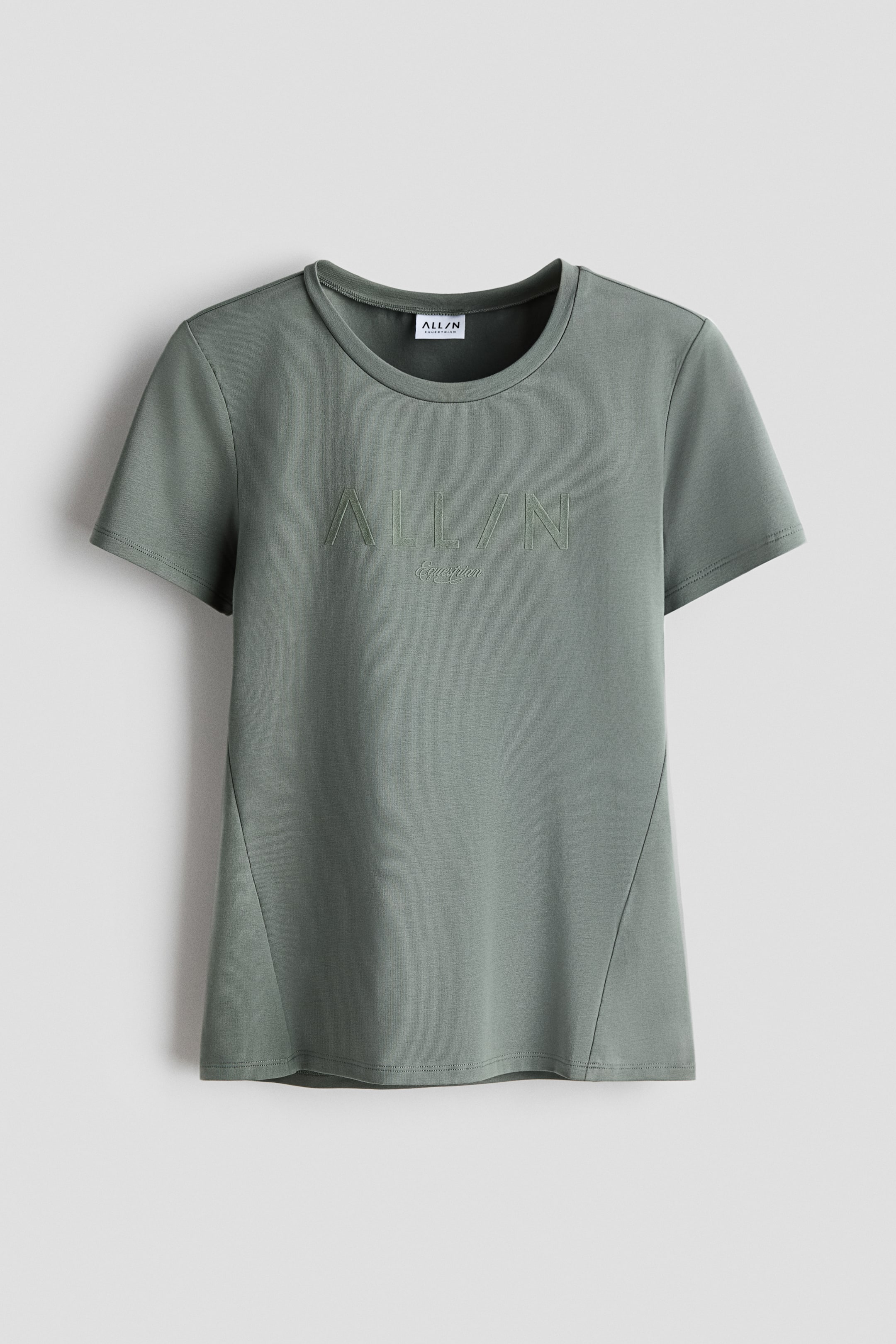View larger image: A sage-green t-shirt is shown from the front, with a crew neck and short sleeves. Embossed 'ALL/N Equestrian' text is centred on the chest, and diagonal seams run from the underarm towards the hem.