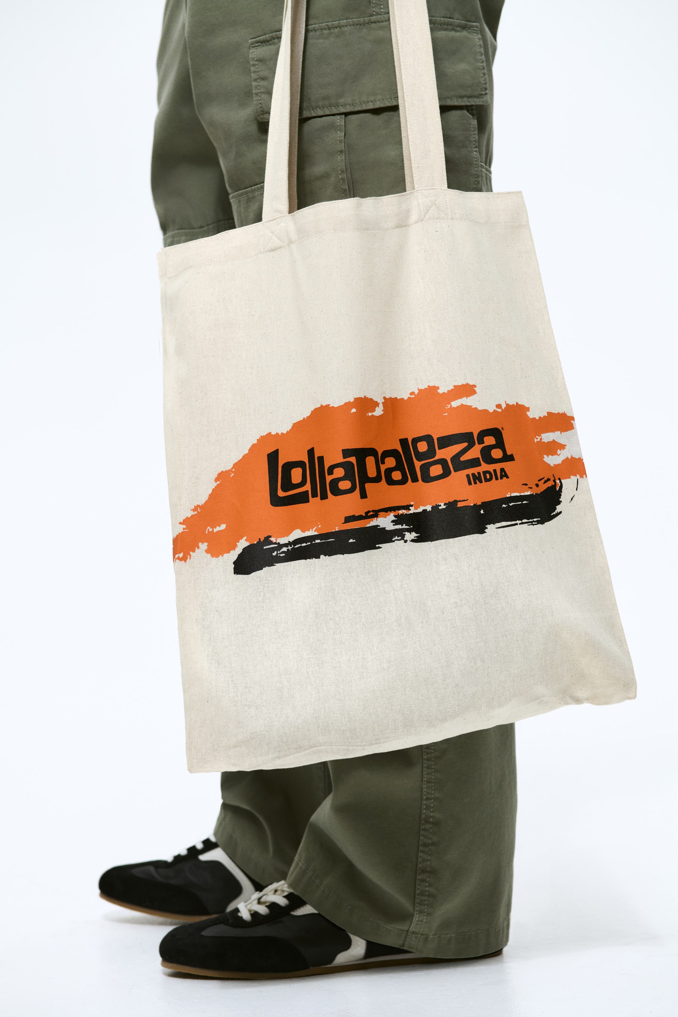 View larger image: A natural canvas tote bag is shown hanging, featuring a bold orange brushstroke graphic with stylised black text “Lollapalooza INDIA” centred on it, along with a black brushstroke detail below. The bag has two fabric handles extending from the top edge.