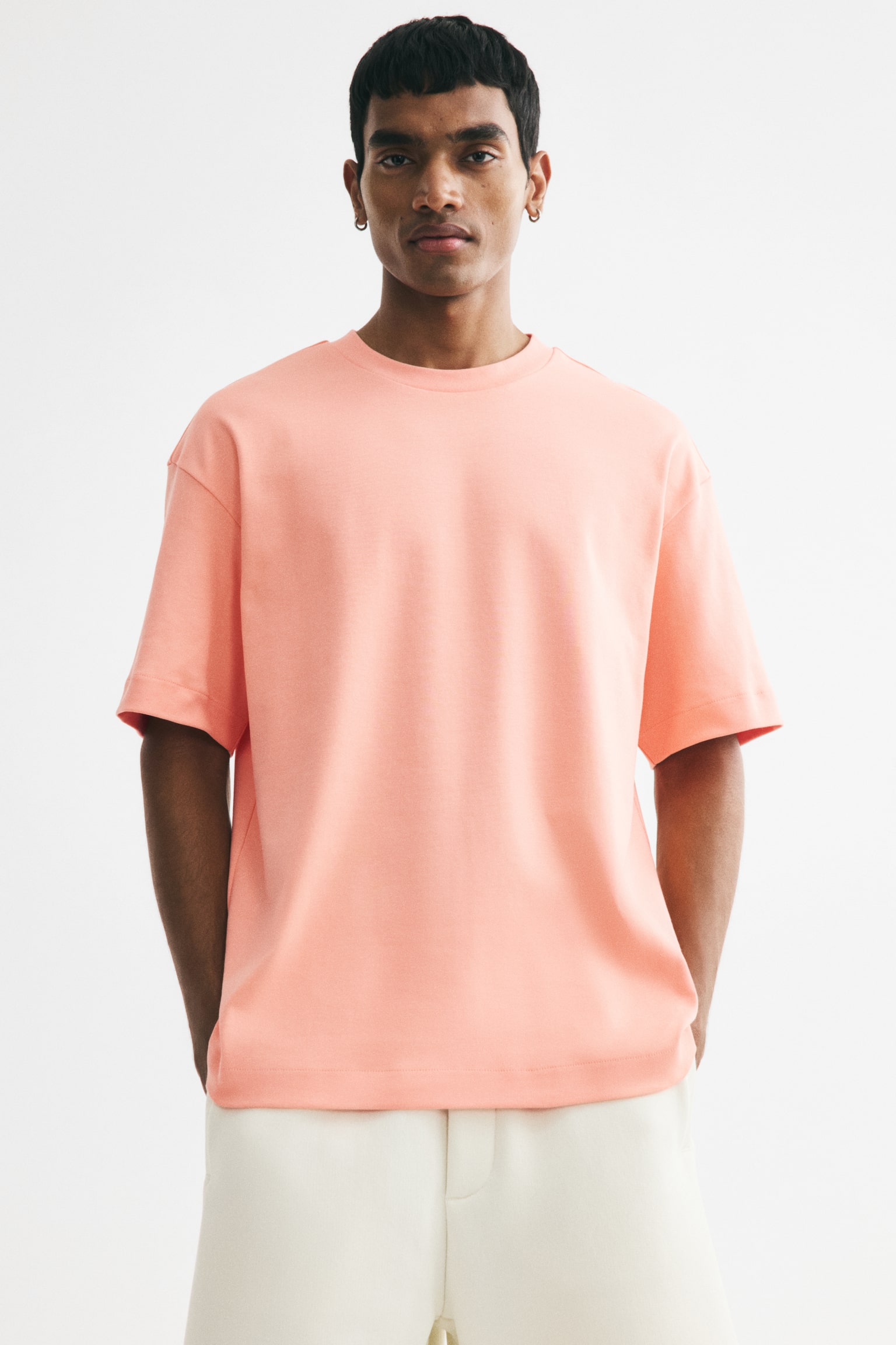 A peach-coloured, boxy-style t-shirt is showcasing its front view, featuring a round, rib-trimmed neckline, dropped shoulders, and a straight-cut hem with a loose fit. - 1