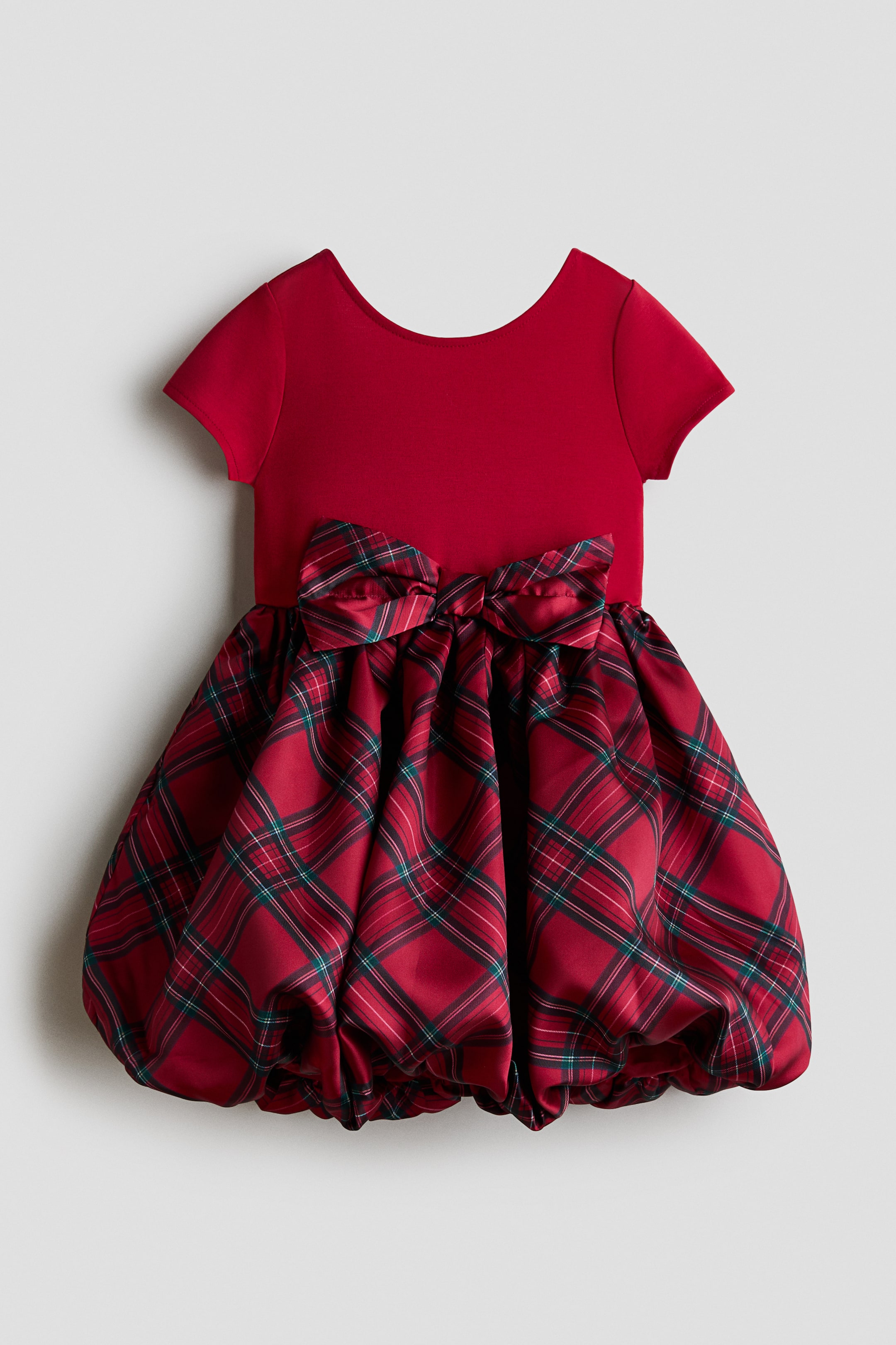 View larger image: A red toddler dress is shown from the front, featuring a solid red knit bodice with short cap sleeves and a scoop neckline. A large plaid bow embellishes the waist, which flows into a voluminous bubble-hem skirt in a red and green tartan plaid pattern.