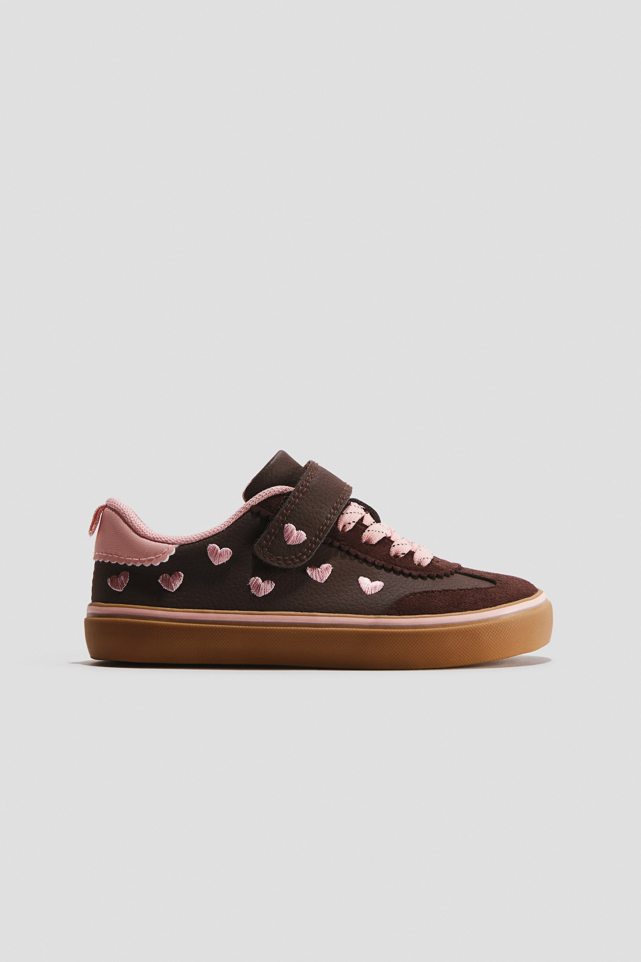 Visualizza immagine più grande: Una sneaker marrone scuro è rivolta a sinistra, caratterizzata da cuori ricamati rosa sulla tomaia. È progettata con un colletto del tallone rosa con bordo smerlato, lacci rosa e un singolo cinturino in velcro marrone, il tutto poggiato su una suola marrone con una striscia rosa.