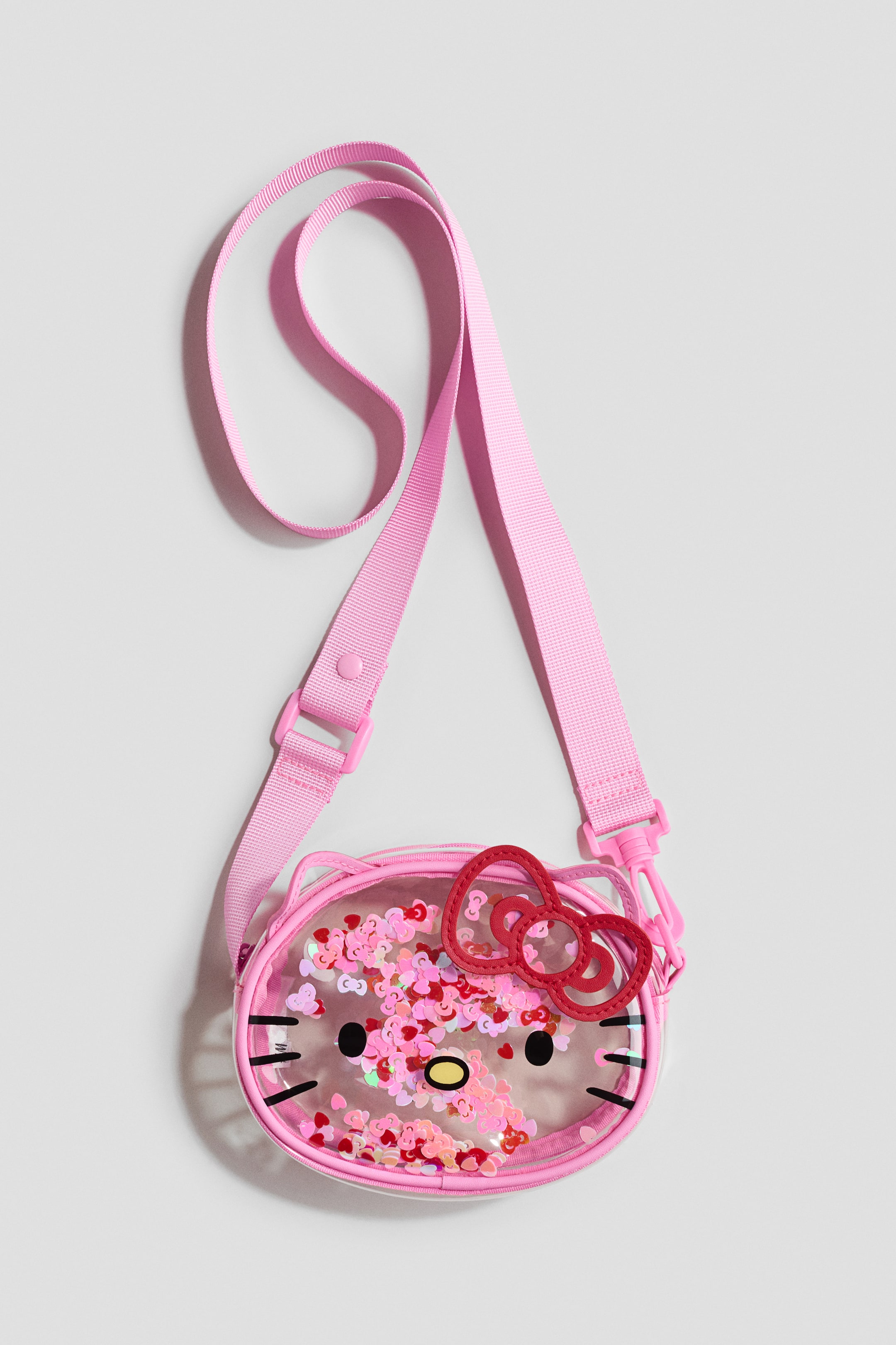 Grotere afbeelding bekijken: Hello Kitty doorzichtige crossbody tas heeft een ronde transparante voorzijde met zwarte Hello Kitty gezichtsomtrekken, inclusief ogen, een gele neus en snorharen. Roze en rode hartvormige pailletten vullen het paneel, en een rode strik van imitatieleer is bevestigd aan de bovenkant rechts. Een roze verstelbare stoffen riem met roze plastic hardware is aan de tas bevestigd.