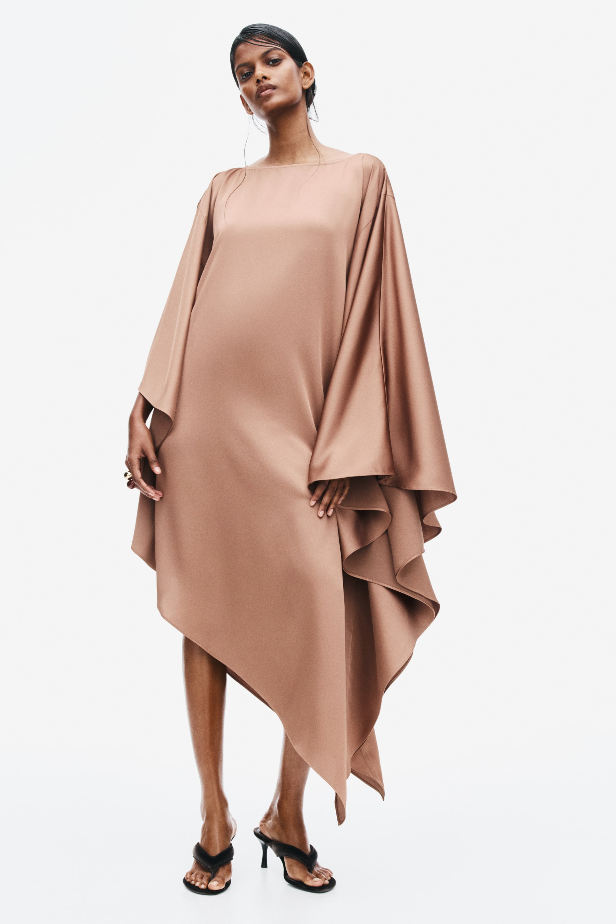 View larger image: The model is wearing a satin cape dress in a taupe shade, featuring a boat neckline, oversized draped cape sleeves, and an asymmetric cascading hemline. The dress is styled with black heeled sandals.