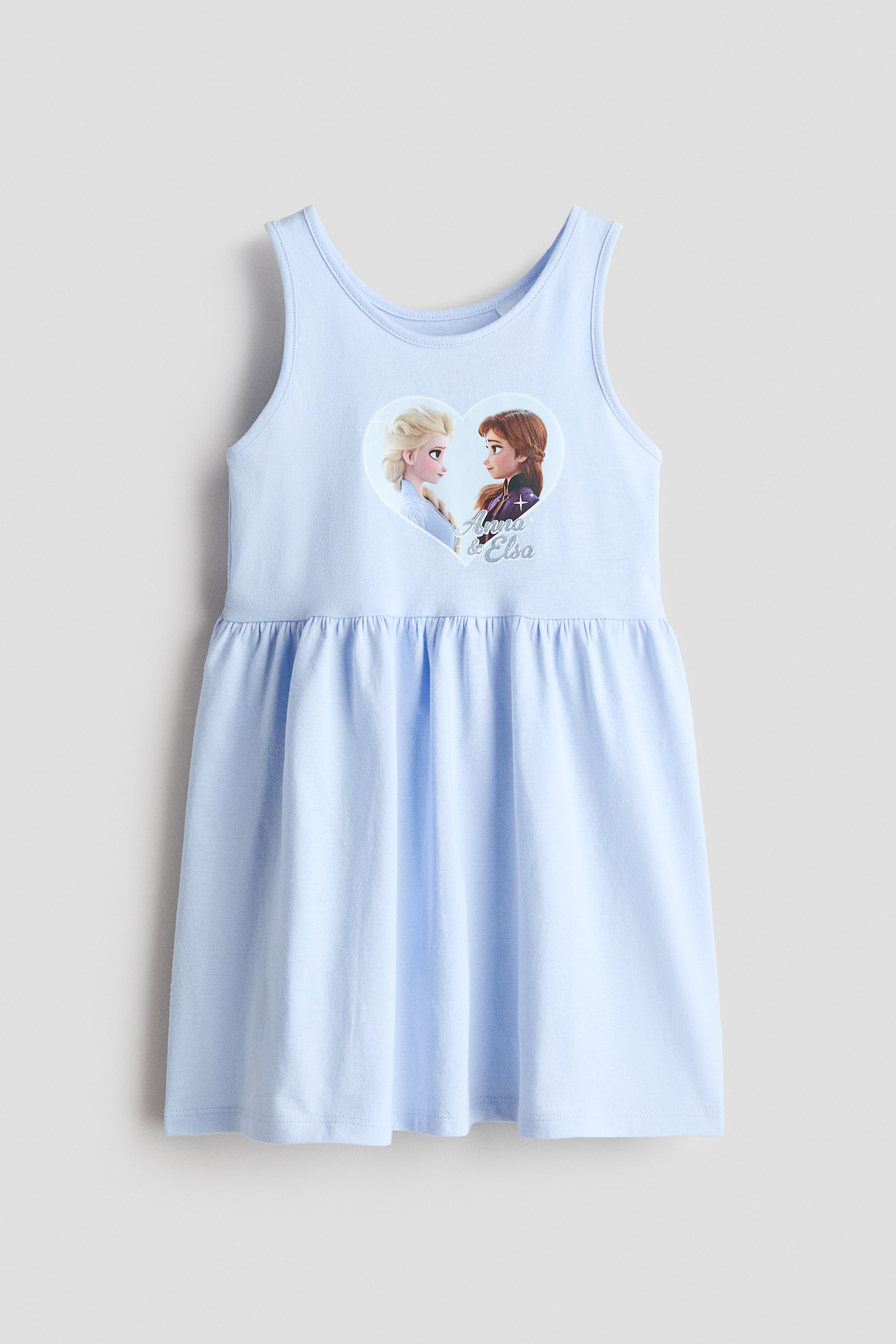 Ingrandisci l'immagine: Il vestito senza maniche, presentato FRONTE, è un design in morbido jersey di cotone azzurro chiaro caratterizzato da uno scollo rotondo con bordi stretti e una grande stampa grafica a forma di cuore di due personaggi femminili con il testo 'Anna & Elsa' sul corpetto. Una cucitura arricciata crea una gonna svasata.
