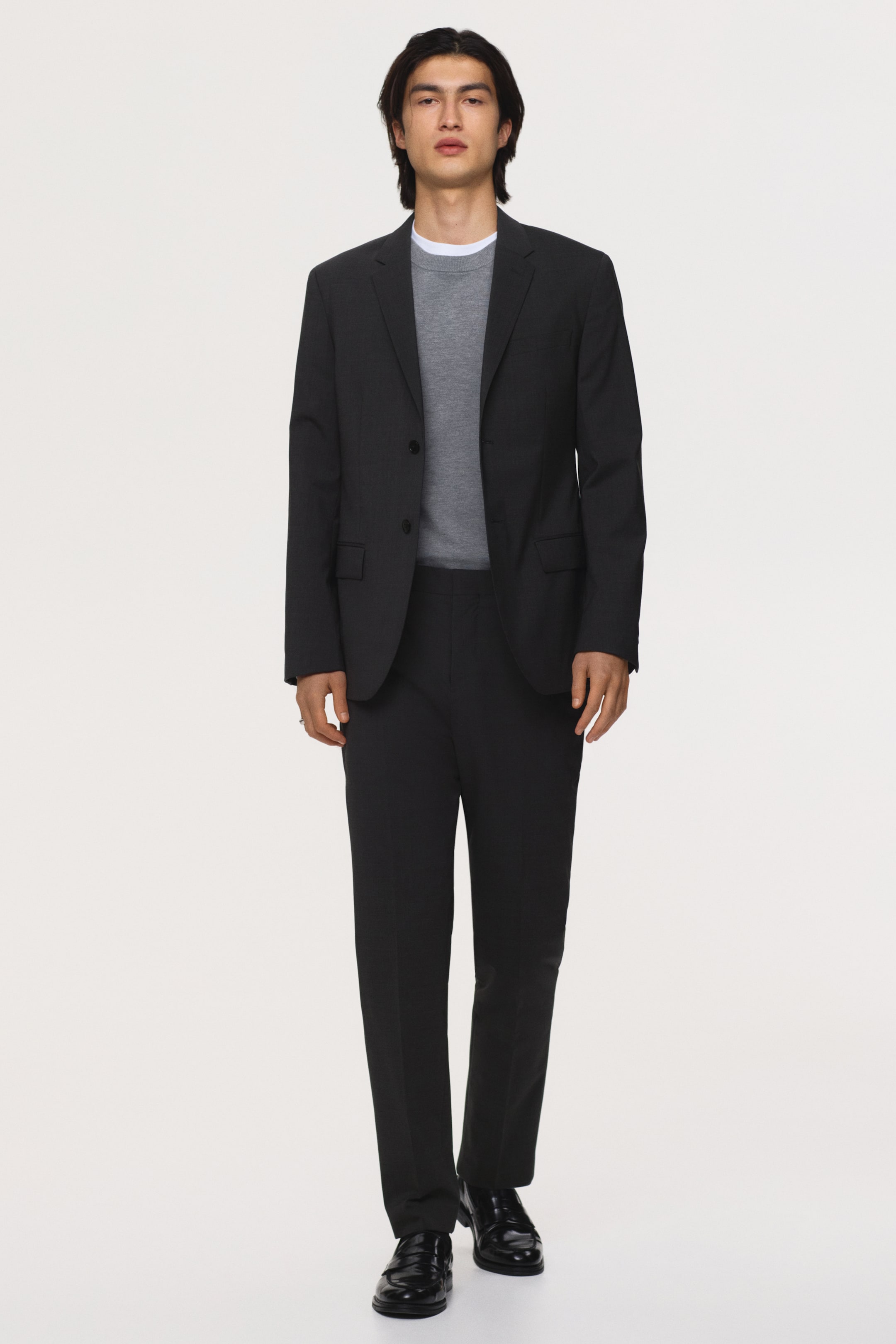 Agrandir l'image: Un costume gris foncé est orienté vers l'avant, comprenant une veste à deux boutons avec des revers crantés et des poches à rabat, ainsi qu'un pantalon droit assorti. Un pull à col rond gris est porté sous la veste, avec le col d'un t-shirt blanc visible, et des mocassins noirs complètent la tenue.