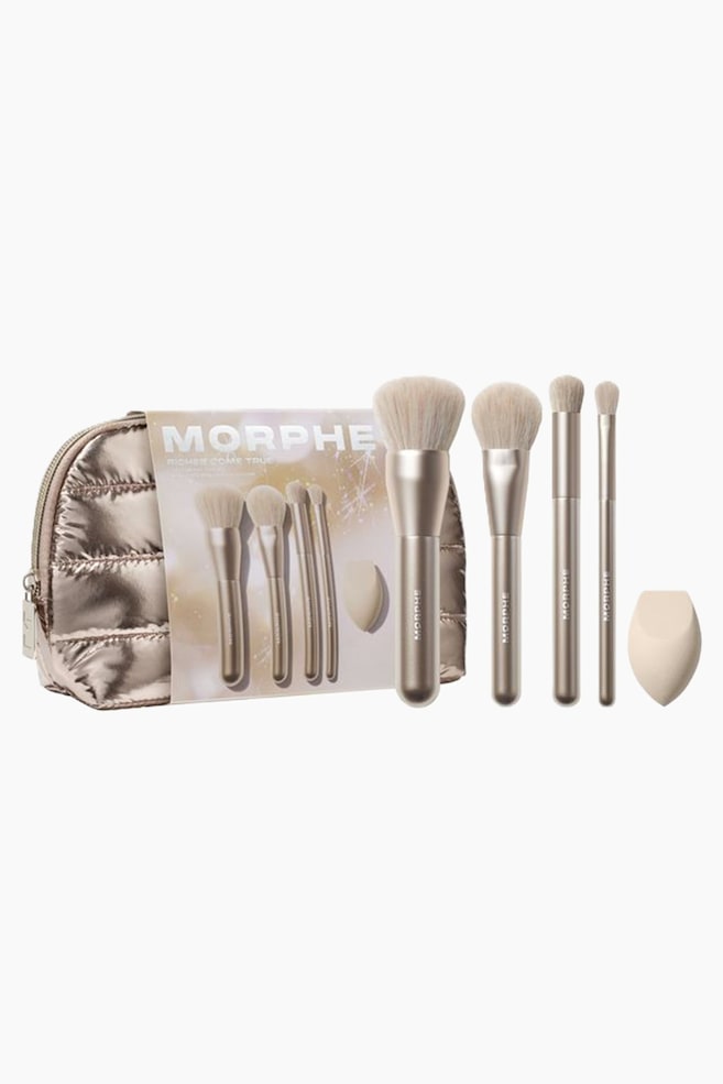 Et femdelt sæt makeupbørster og redskaber fremviser fire champagneguld-metalliske børster med lyse beige børstehår sammen med en dråbeformet beige makeupsvamp, der står oprejst. En quiltet makeup-taske i metallisk bronze med lynlås står til venstre bag produktets brandede emballagekort. - 1