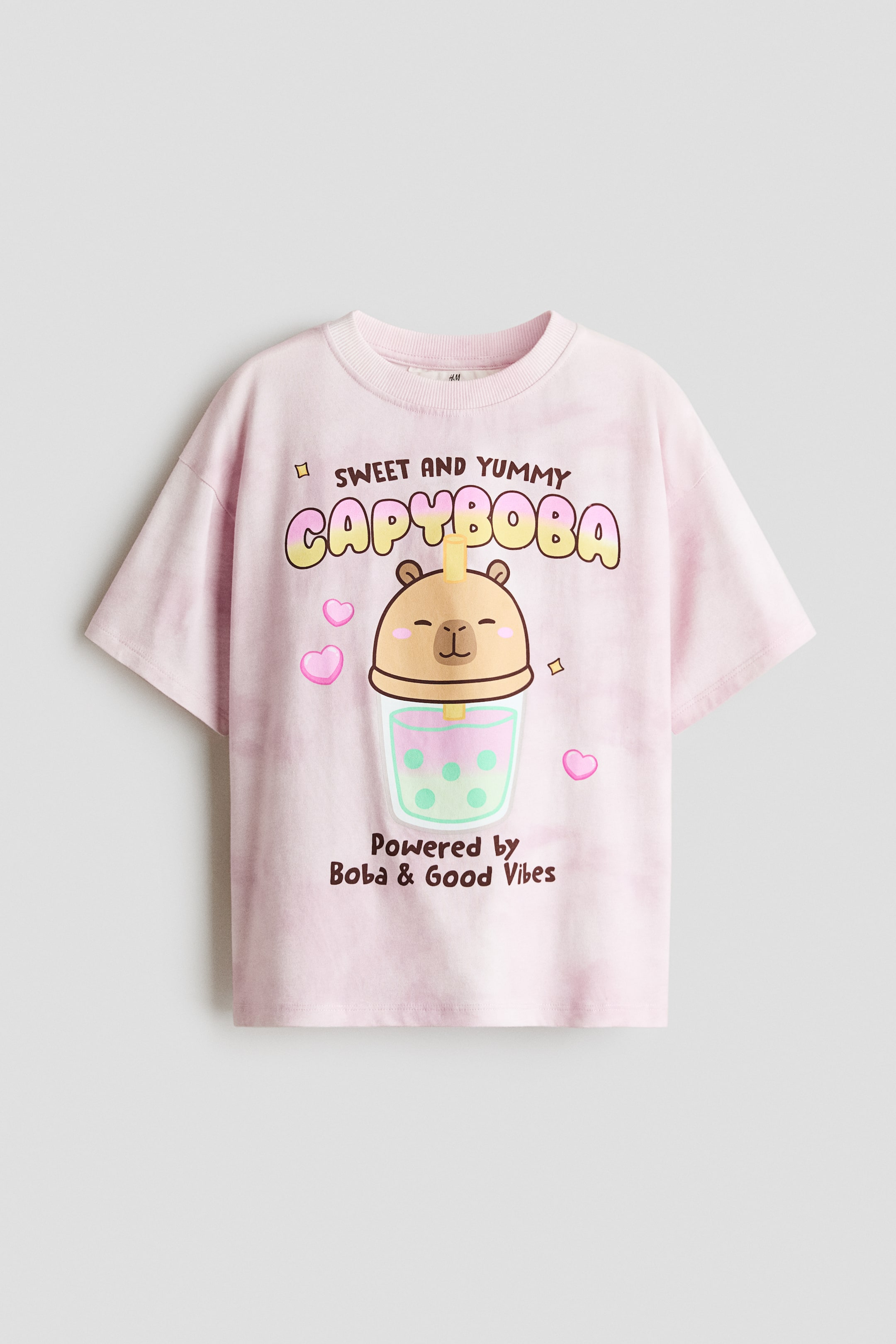 Grotere afbeelding bekijken: Een lichtroze T-shirt toont zijn voorkant, met een gemêleerde katoenen jersey stof, een ronde halslijn met ribboord en verlaagde schouders. Op de voorkant siert een grote grafische print, die een cartoon capibara-hoofd op een boba-theekopje afbeeldt, omringd door roze hartjes en tekst met de woorden 'SWEET AND YUMMY CAPYBOBA' en 'Powered by Boba & Good Vibes'.
