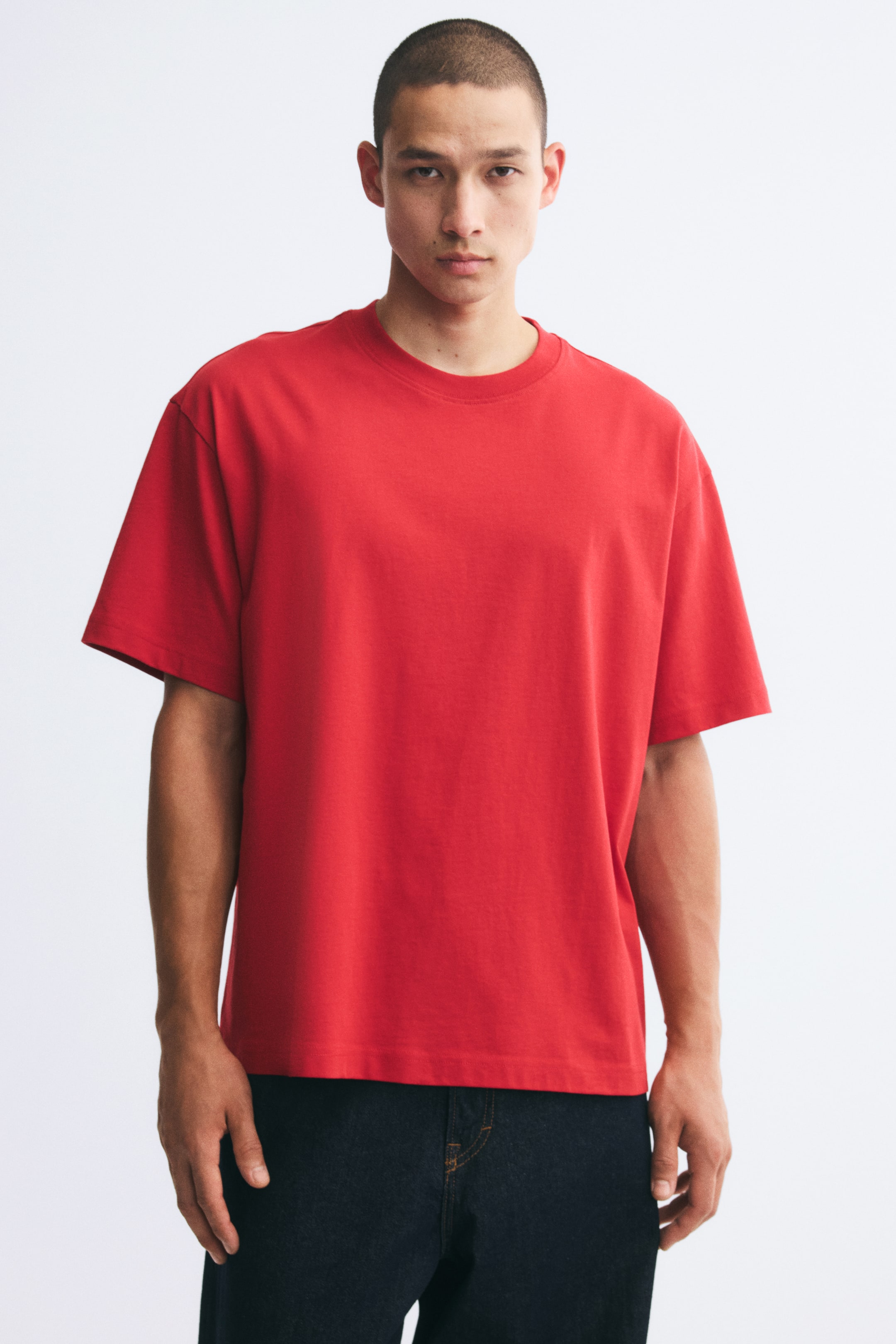View larger image: A red cotton jersey T-shirt featuring a relaxed fit, a round rib-trimmed neckline, and dropped shoulders is being worn by the model. The T-shirt has a straight-cut hem and is styled with dark denim jeans.
