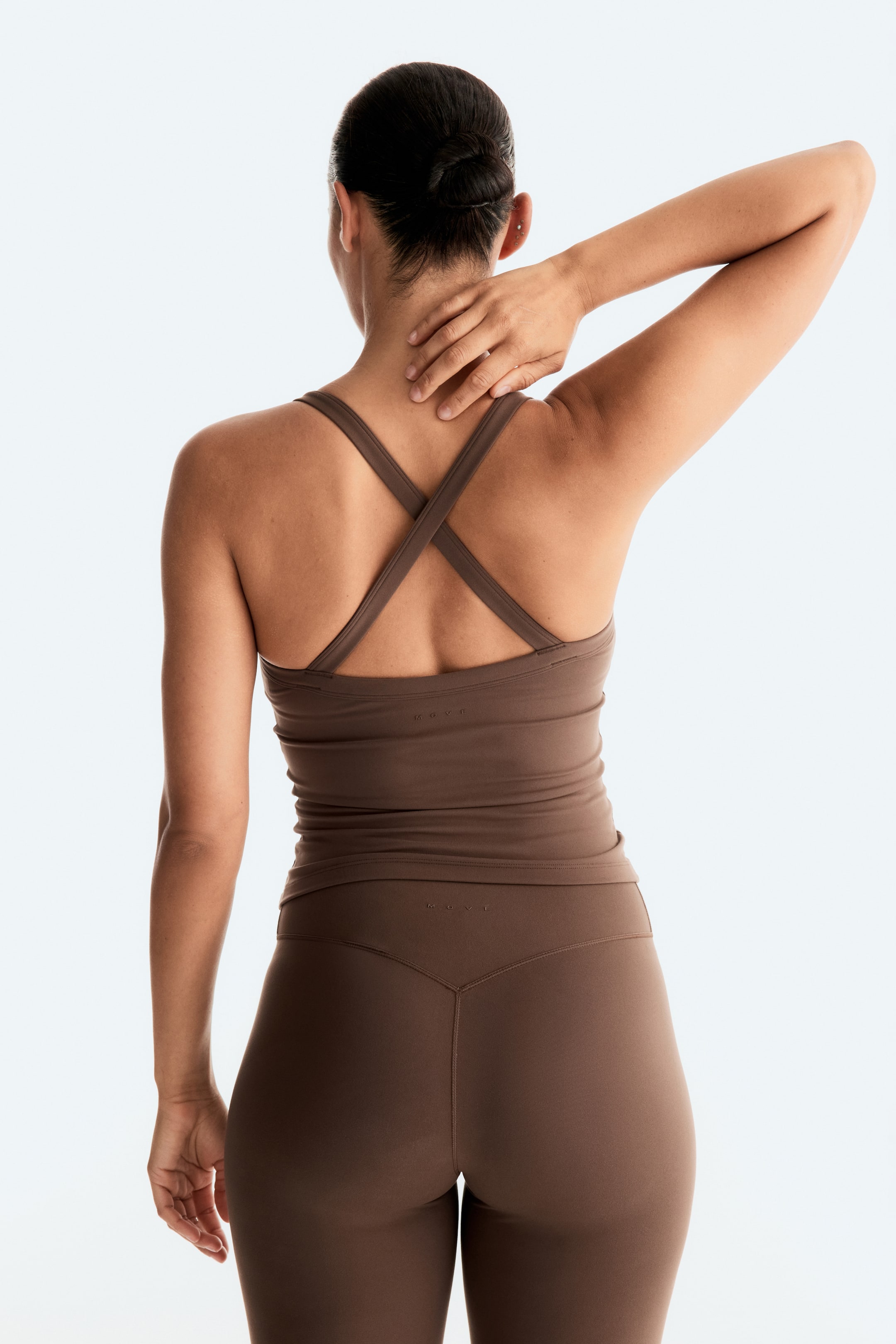 View larger image: A brown activewear set is showcased from the back, highlighting a fitted tank top with criss-cross back straps and an embossed 'MOVE' logo visible at mid-back. The matching high-waisted leggings feature a V-shaped seam detail and an embossed 'MOVE' logo on the back waistband.