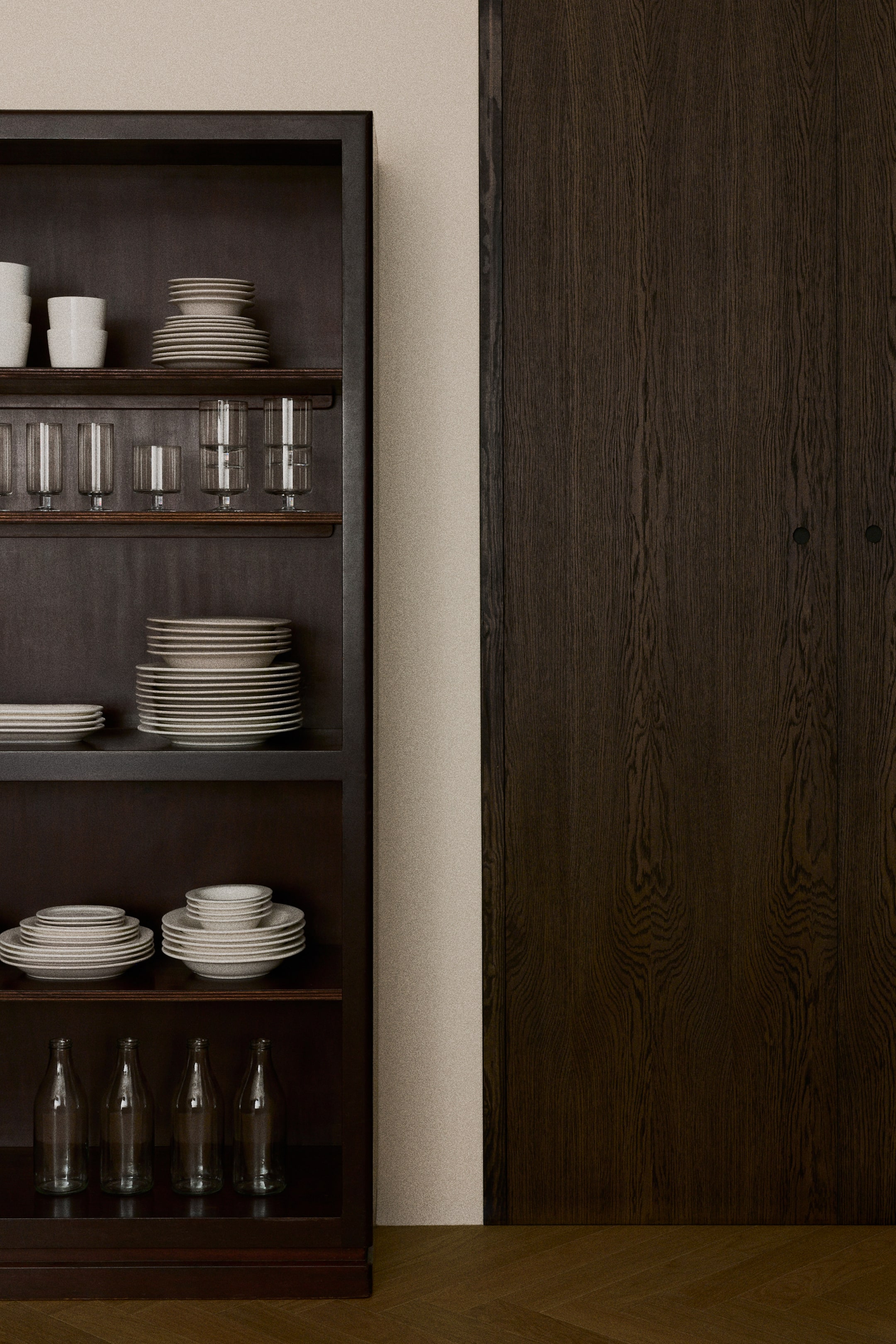 View larger image: A dark-stained wooden shelving unit is shown in a front-facing view, displaying neatly arranged rows of neutral-toned ceramic tableware and glassware on its open shelves. The shelves hold stacks of textured beige plates and bowls, white ceramic mugs, clear tumblers, and glass bottles.