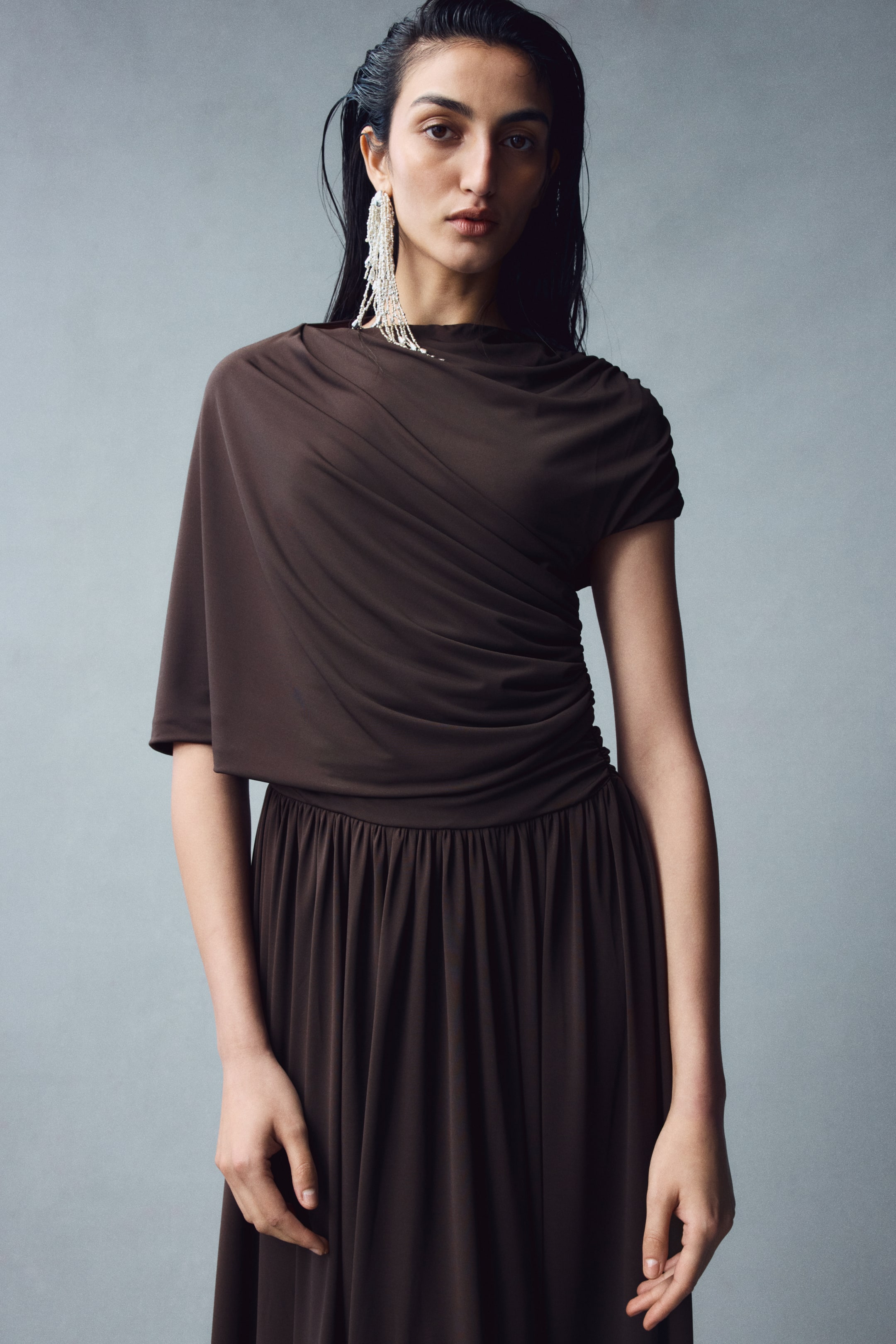 View larger image: The model is wearing a dark brown stretch jersey dress featuring a high neckline, an asymmetrical draped sleeve panel on one side, and a short sleeve on the other. The dress has a fitted bodice with gathered draping throughout, a defined gathered waist, and a flared skirt.