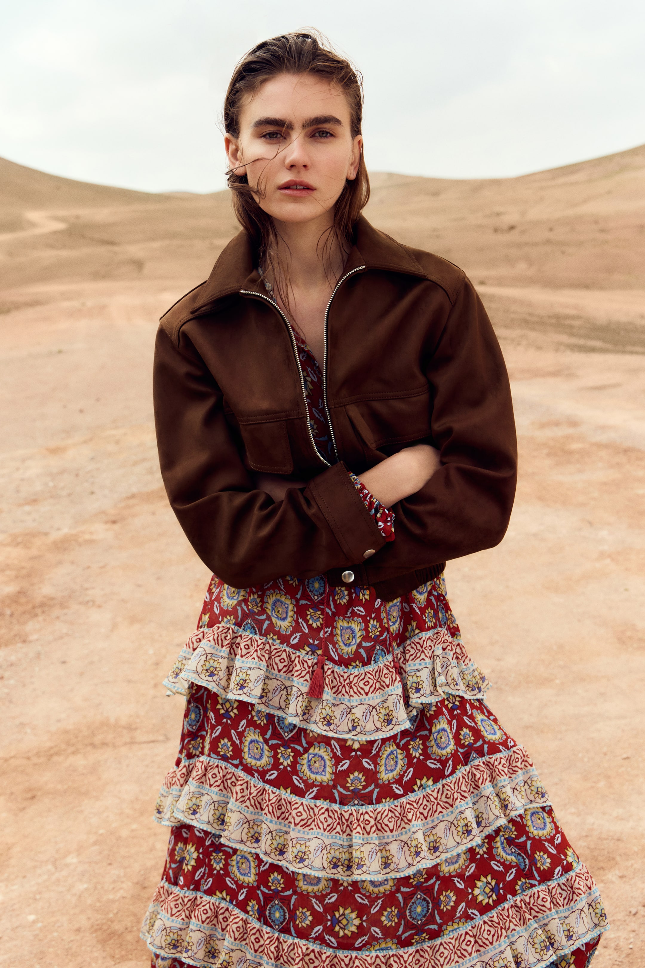 View larger image: The model is wearing a brown coated fabric jacket, showcasing a collar, diagonal shoulder seams, flap front pockets, and an elastic hem. The front zipper of the jacket is partially open, and the model has their arms crossed over the jacket, which is layered over a red patterned dress.
