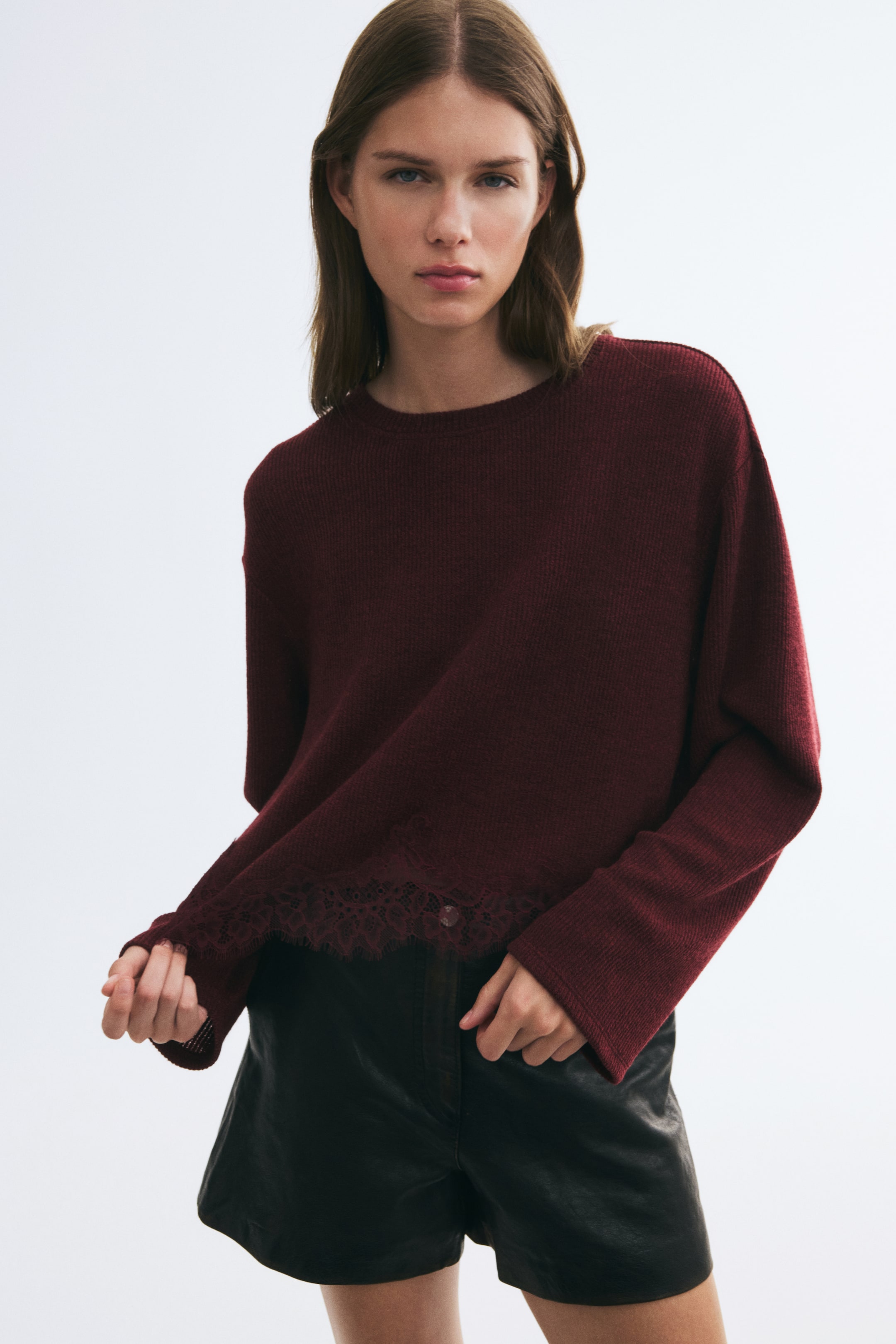 Grösseres Bild ansehen: Ein weinrotes Stricktop mit Rundhalsausschnitt, überschnittenen Schultern und einem feinen, mit Spitze verzierten Saum wird vom Model getragen. Das Model hält mit einer Hand den Spitzenrand des Tops und trägt dazu schwarze Leder-Shorts.