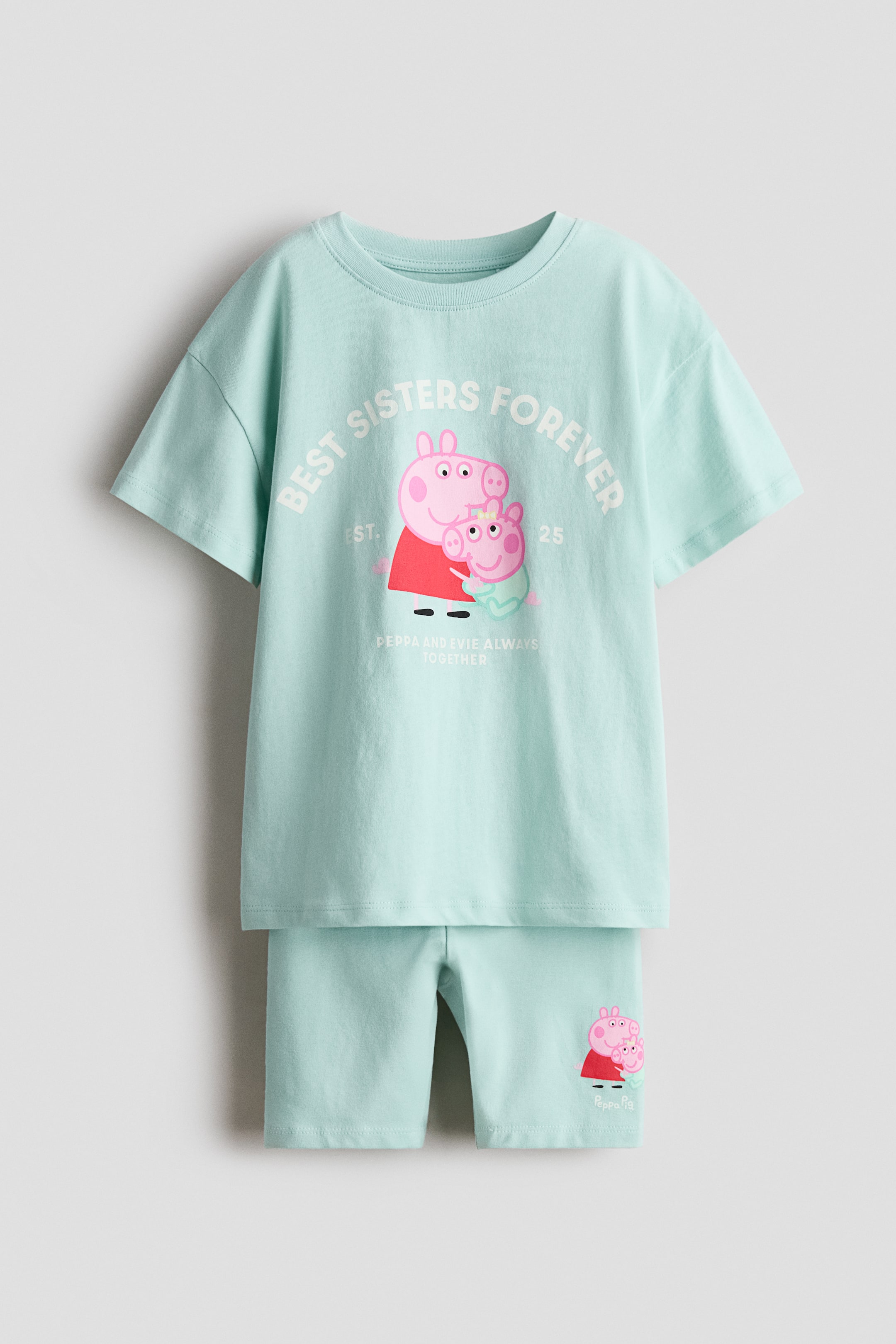 View larger image: The printed set features a light blue T-shirt and matching cycling shorts, both crafted from soft cotton jersey, showcased from the front. The T-shirt boasts a ribbed trim around the neckline, dropped shoulders, and a central graphic of two pink pig characters with the text "BEST SISTERS FOREVER" and "PEPPA AND EVIE ALWAYS TOGETHER." The cycling shorts display an elasticated waist and a smaller matching pig graphic on the left leg.