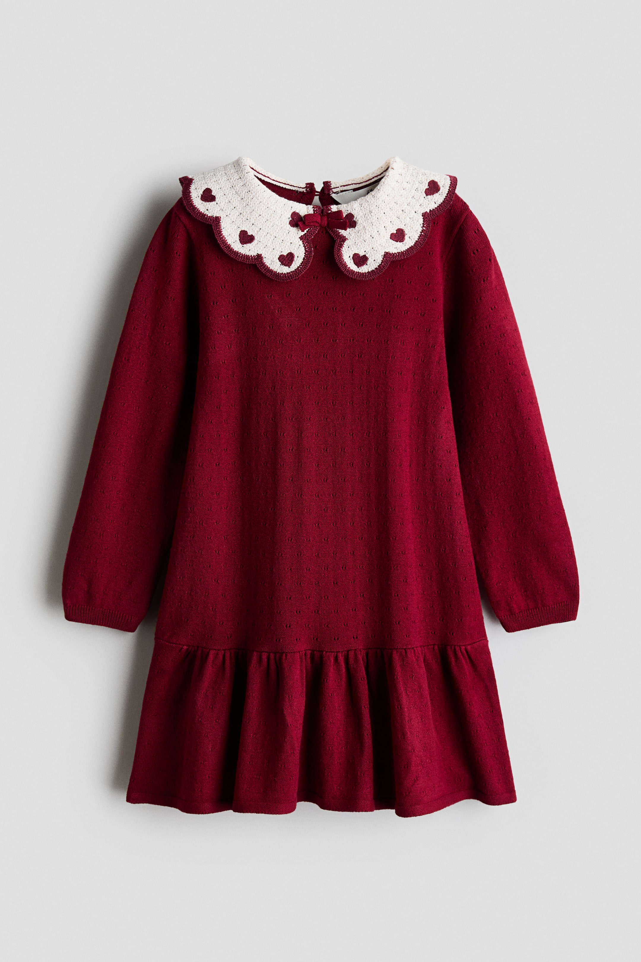 View larger image: A long-sleeved, deep red knit dress is shown facing front, featuring a scalloped white crochet Peter Pan collar detailed with embroidered red hearts and a small bow at the centre. The dress has a textured pointelle knit body, ribbed cuffs, and a flared, ruffled hem.