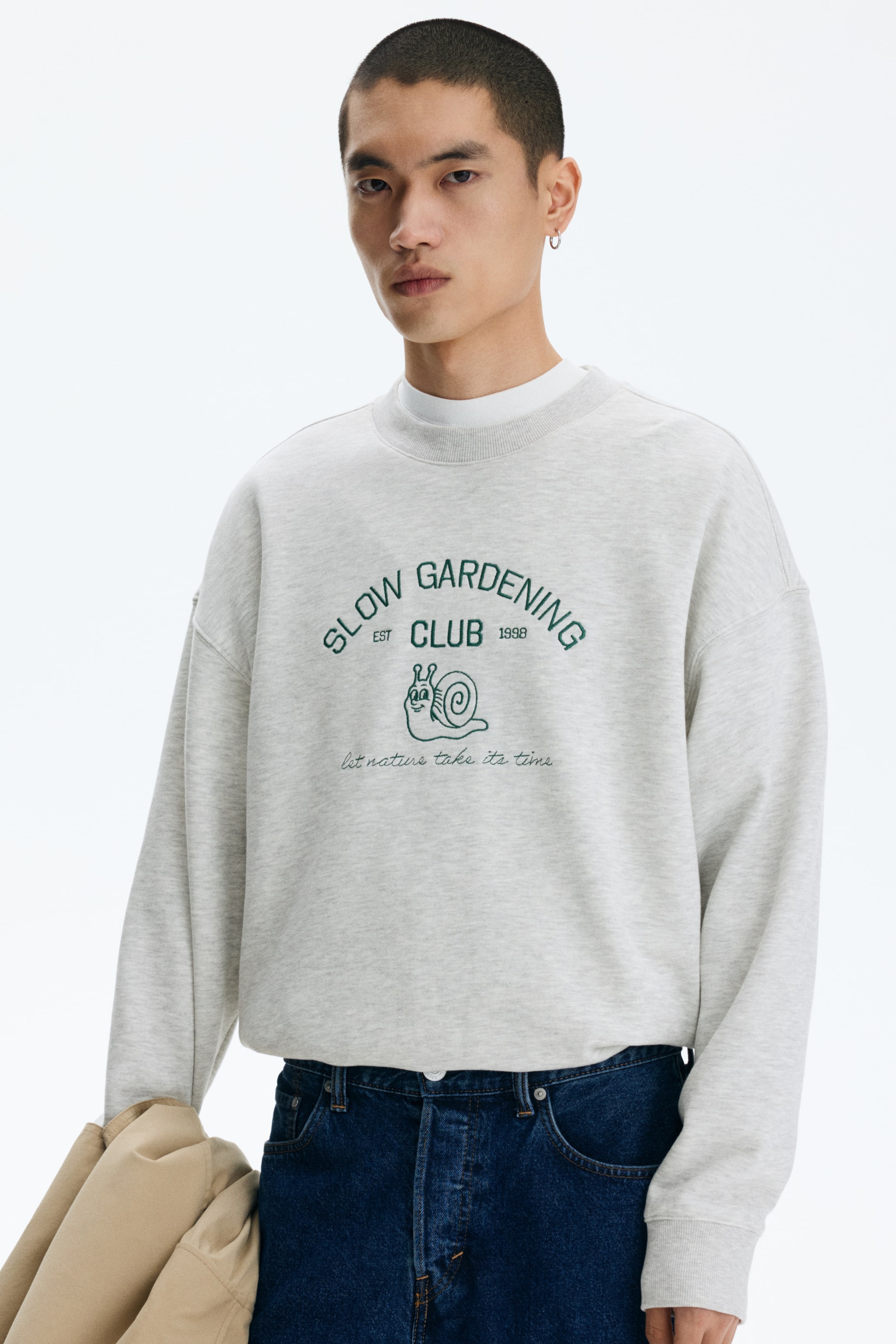 이미지 확대 보기: 연한 회색 멜란지 크루넥 스웨트셔츠를 착용하고 있으며, 앞면에는 녹색 "SLOW GARDENING CLUB EST 1998" 그래픽과 만화 달팽이, "let nature take its time" 문구가 표시되어 있습니다. 이 스웨트셔츠는 긴 소매에 리브드 커프스와 리브드 밑단이 특징이며, 흰색 티셔츠 위에 레이어드되어 있습니다.
