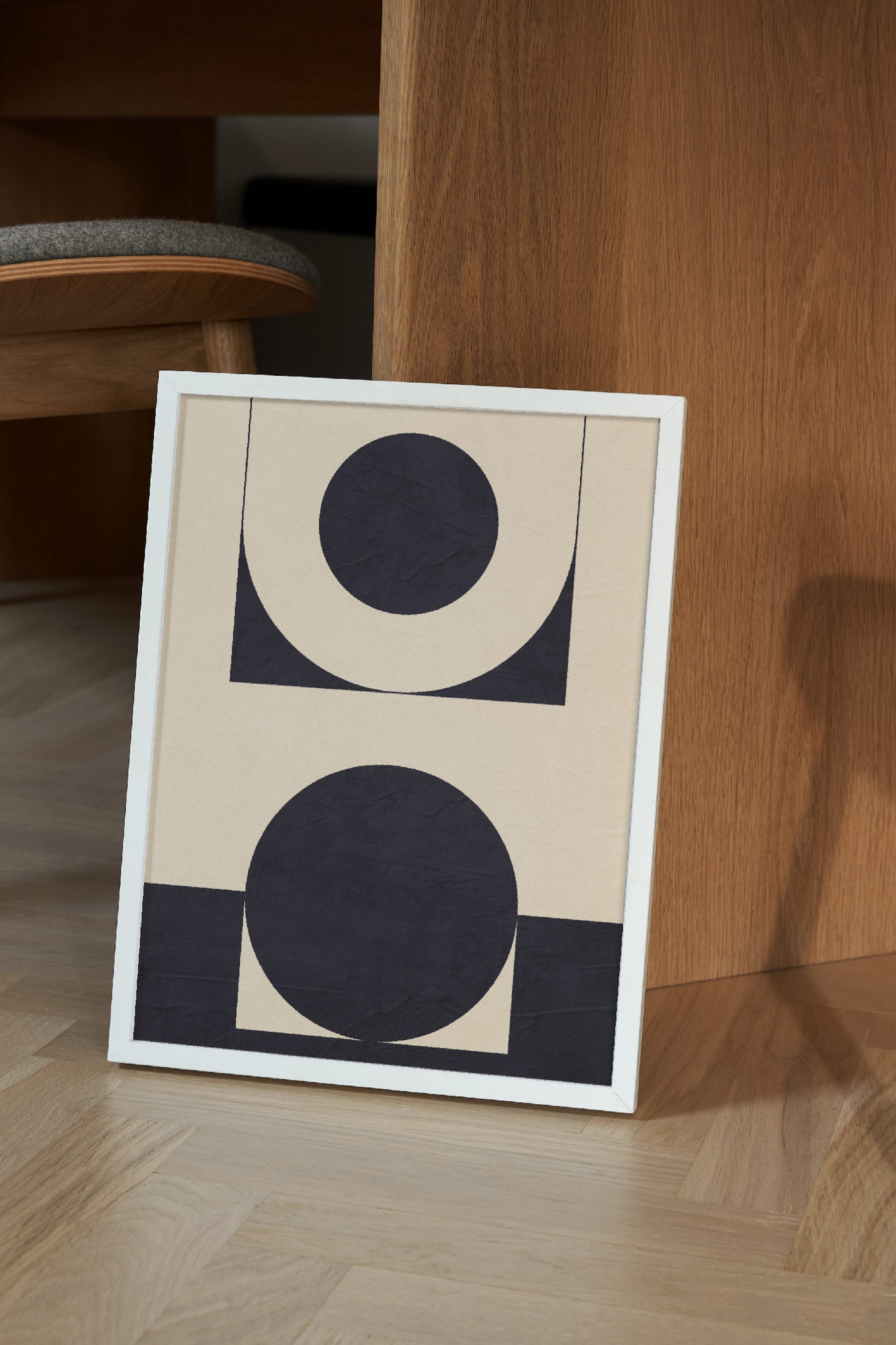 View larger image: A white-framed artwork is leaning, showcasing an abstract design with a beige background. The design features two large, dark, textured circles, each accompanied by a dark quarter-square shape in opposite corners of their respective halves, and thin dark lines.