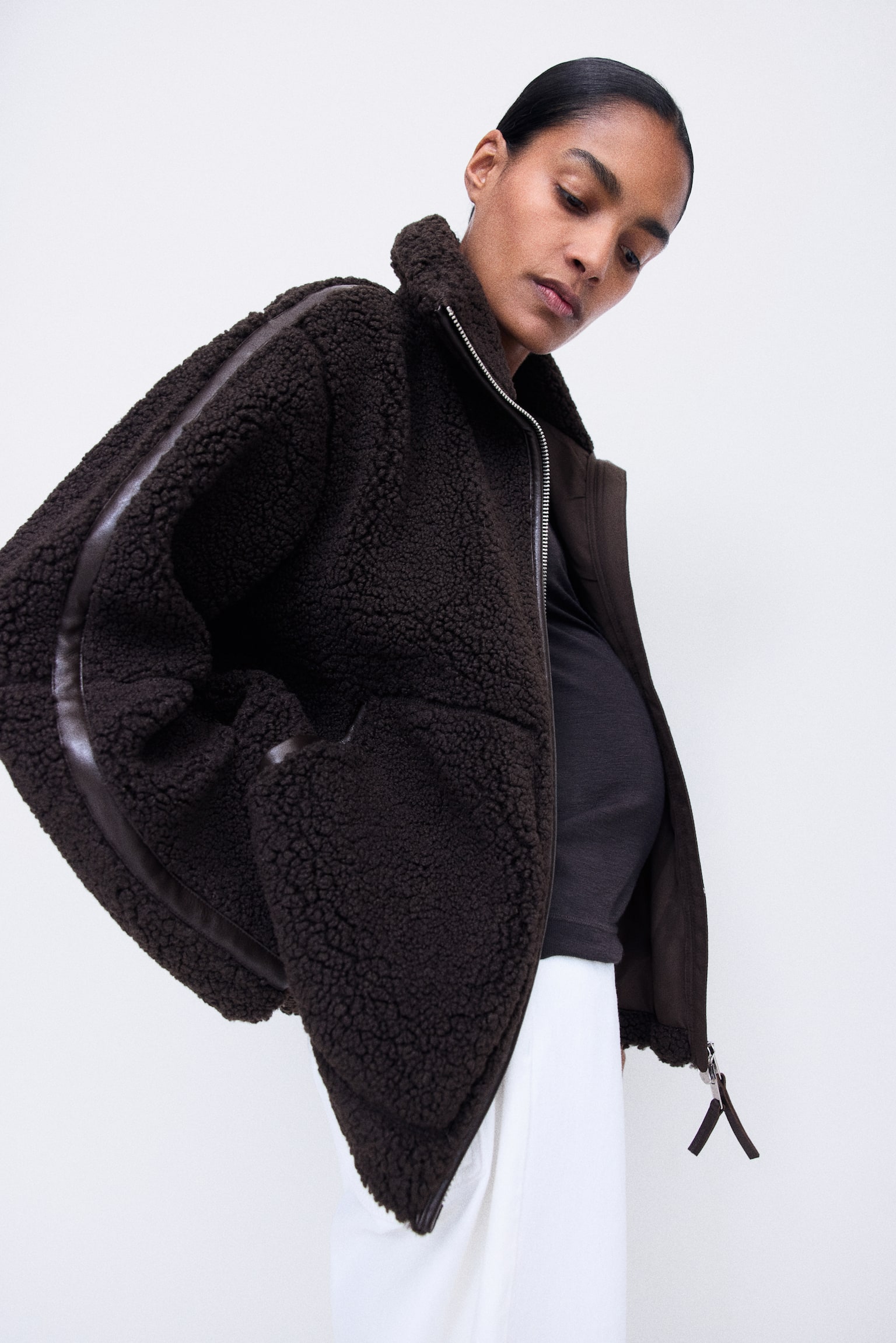 Eine dunkelbraune Teddy-Fleece-Jacke wird von vorne gezeigt, mit lockerer Passform, hohem Stehkragen und silberfarbenem 2-Wege-FrontreiÃverschluss mit dunkel beschichteter Einfassung. Passend dazu verlÃ¤uft eine beschichtete Einfassung entlang der Schultern, und groÃe aufgesetzte Taschen sind beidseitig auf der Vorderseite platziert. - 1